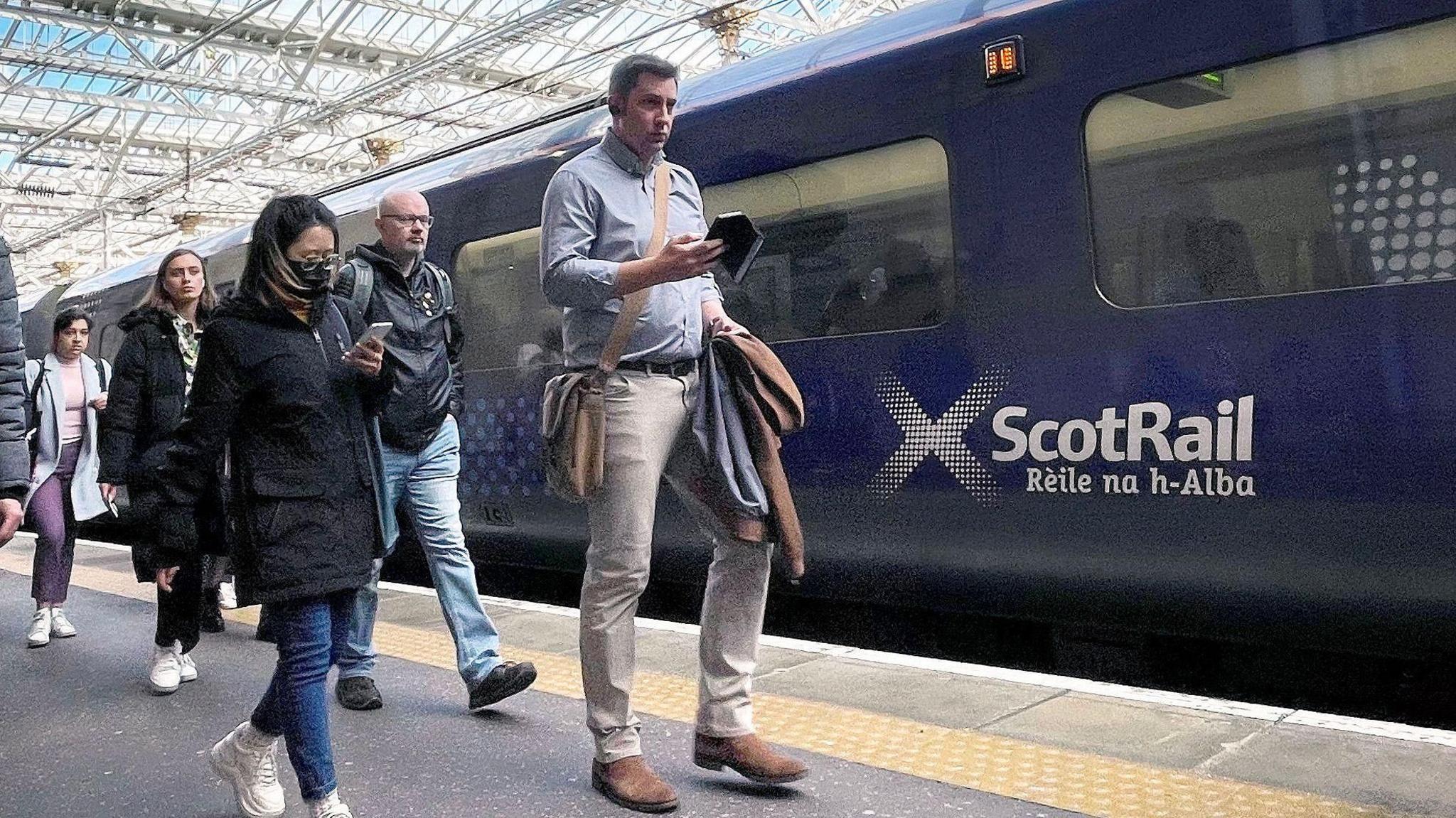 Group of train passengers walking on platform, a man with a bag looks at his phone, with blue scotrail train at station under a glass roof canopy