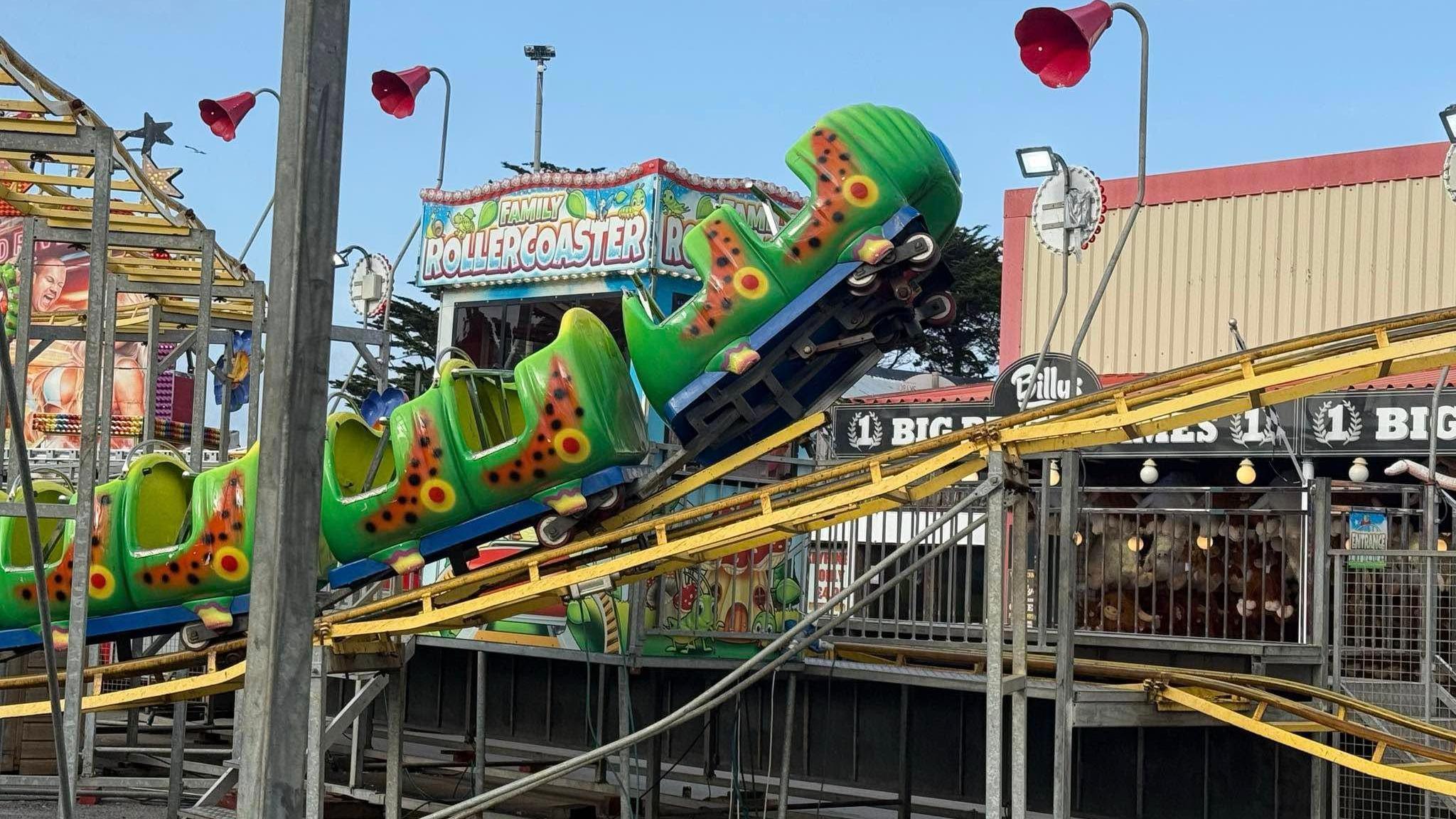 A side on view of a caterpillar themed rollercoaster where the front of the ride has come off the tracks. It is suspended in the air at the front as the yellow track goes out of the image. Behind there is a sign for a family rollercoaster