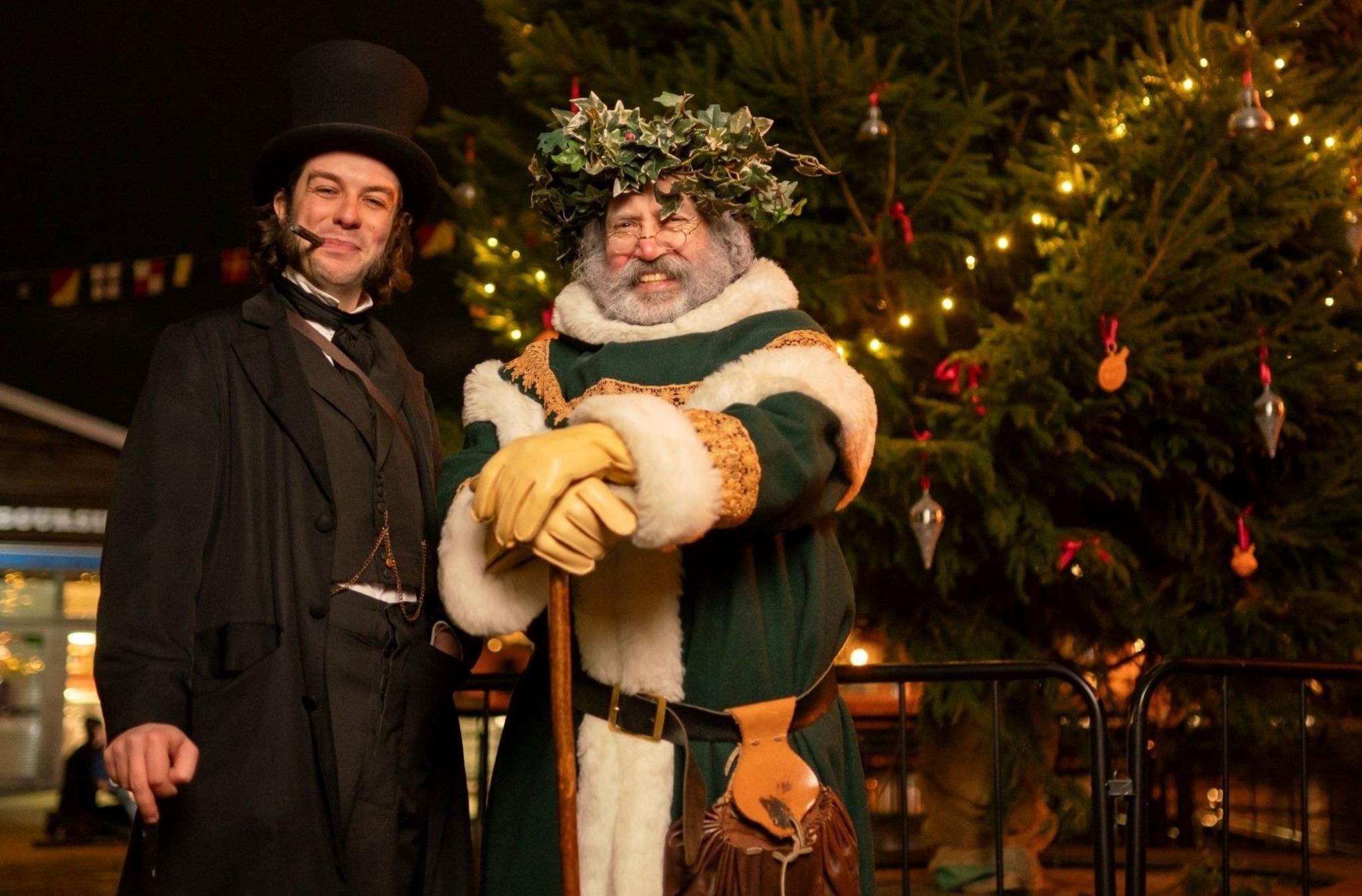 Two actors, one dressed as Isambard Kingdom Brunel and another as Father Christmas in a green outfit with white fur trim, stand next to a Christmas tree at the SS Great Britain in Bristol. They are both smiling at the camera and the picture is taken at night, with the tree's lights on