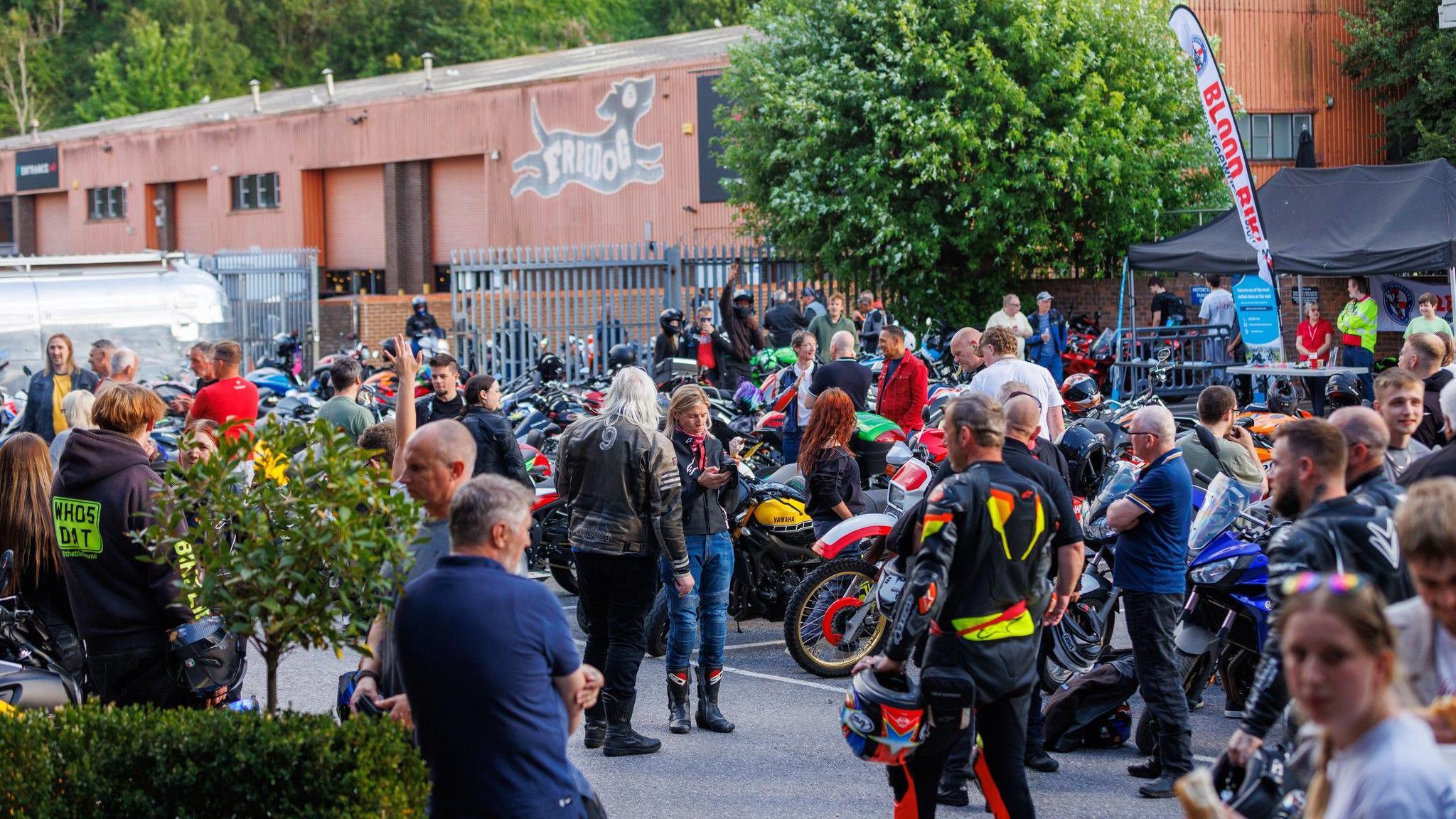 Dozens of people stand around motorbikes in the main car park at Fowlers Motorcycles in Bristol. Many of them are wearing biker leathers. In the background the side of activity centre Freedog is visible.