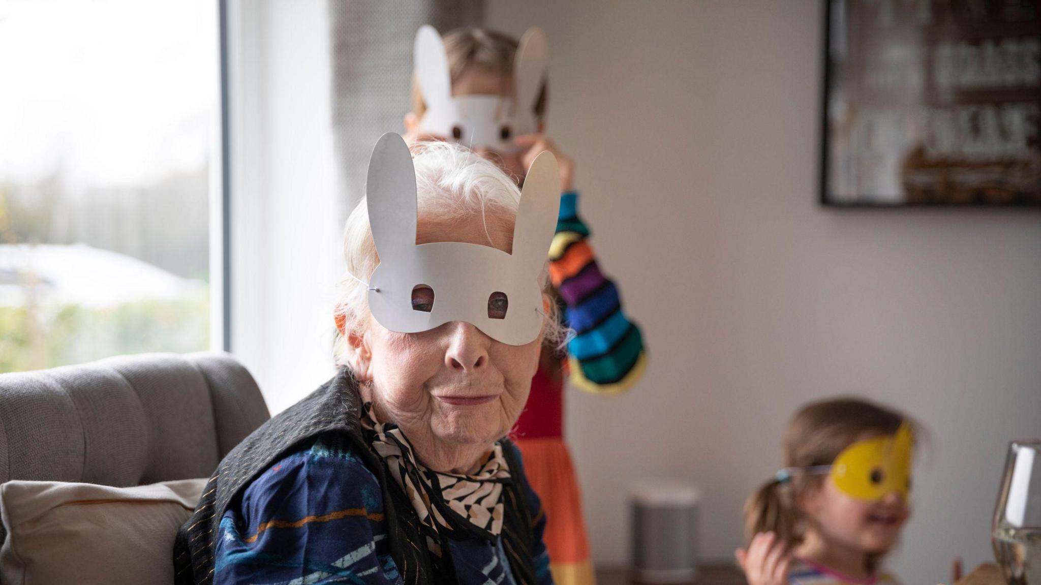 Grandmother wearing a novelty Easter bunny mask and pulling a pouting facial expression whilst sitting for lunch at a dining table, with two happy grandchildren wearing novelty masks in the background.