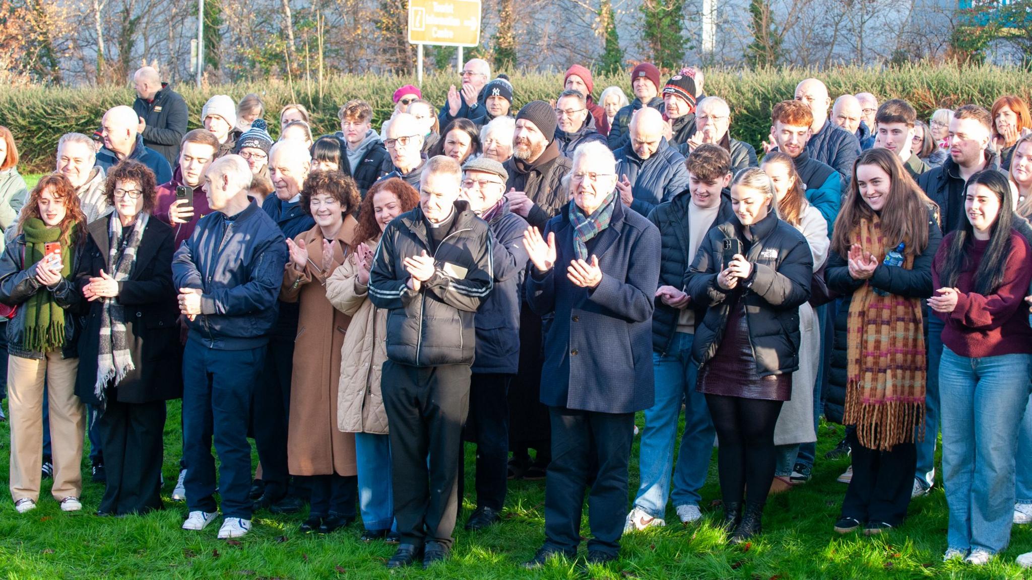 A large number of people, many of whom are dressed in coats and scarves, are standing on a grassy bank, pictured applauding during the ceremony on Saturday.