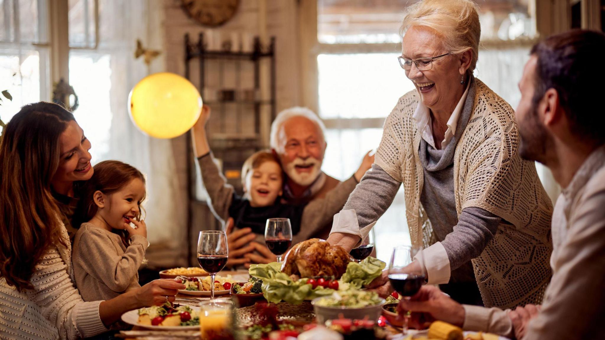 Family around the table enjoying a Christmas dinner. Everyone is smiling, as an older woman lays a turkey on the table. 