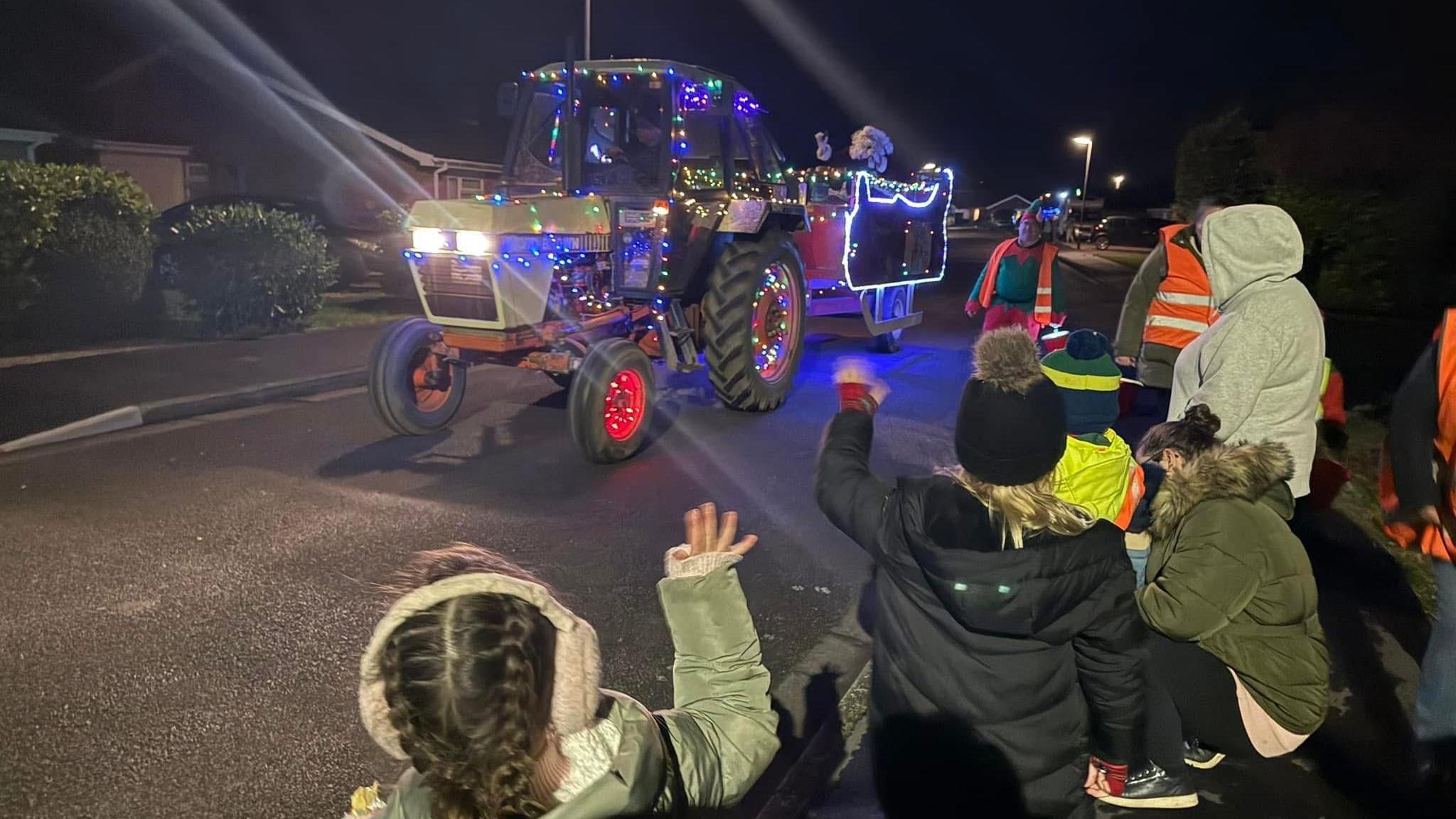 A group of children and adults waving at a colourfully decorated grey vintage tractor festooned with fairy lights and pulling a trailer that looks like a sledge containing Santa dressed in traditional red costume with white beard and gloves waving back at the public.