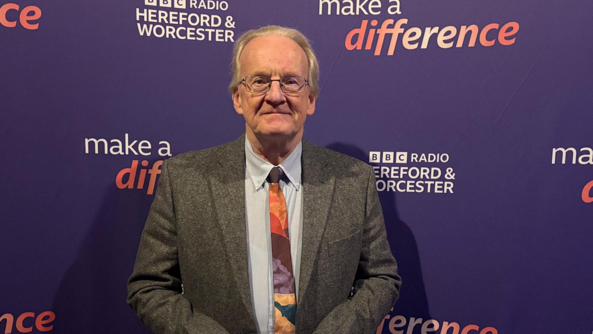 An elderly man in a grey tweed suit, light blue shirt and a multicoloured tie. He has grey receding hair, thin rimmed glasses and is smiling at the camera