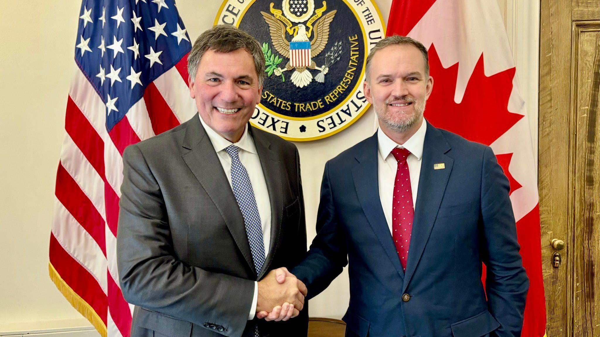 An image showing US trade representative Jamieson Greer (right) shaking hands with Canada-US trade minister Dominic LeBlanc to his left. Behind them are a US and Canada flag with the seal for the US trade representative in the middle. Both men are wearing suits and smiling at the camera.
