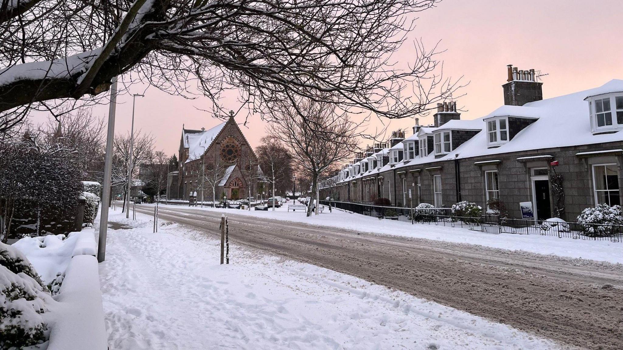 A picturesque scene in Aberdeen. Thick snow is on the ground. There are lots of footprints walking through it on the path. There is a road still covered in slush that has now turned brown. A row of houses have white roofs and an old building stands in the background. There is a pinky/yellow sky.