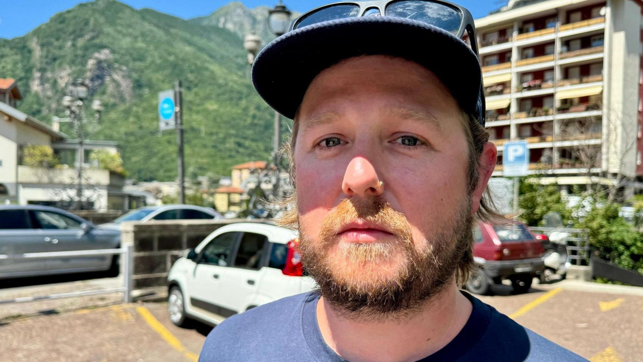 Head and shoulders shot of a man with a short beard wearing a cap with sunglasses perched on the rim. There is a tall white building and mountains in the background.
