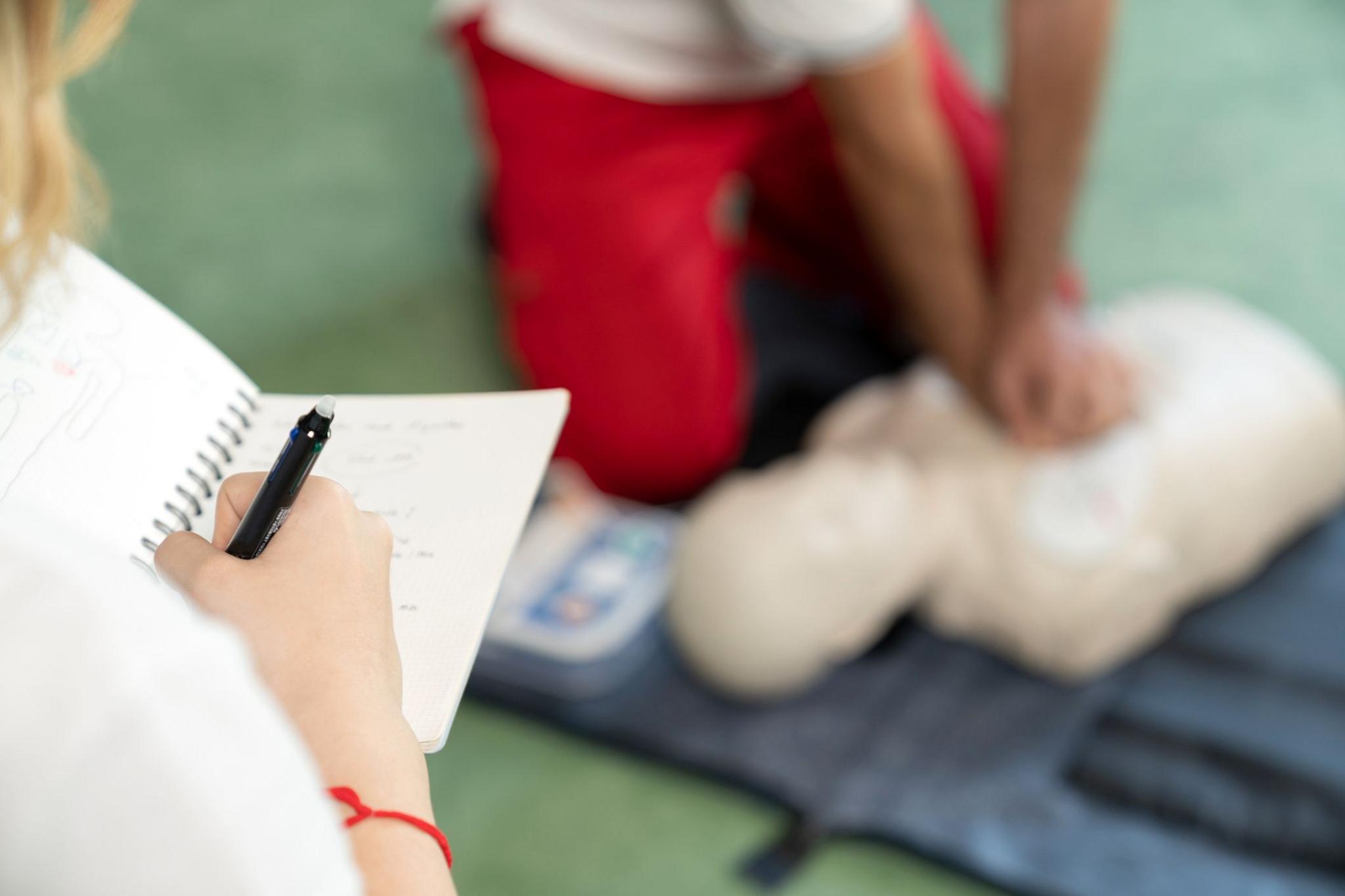 A young person demonstrates CPR skills in a classroom while someone else writes notes