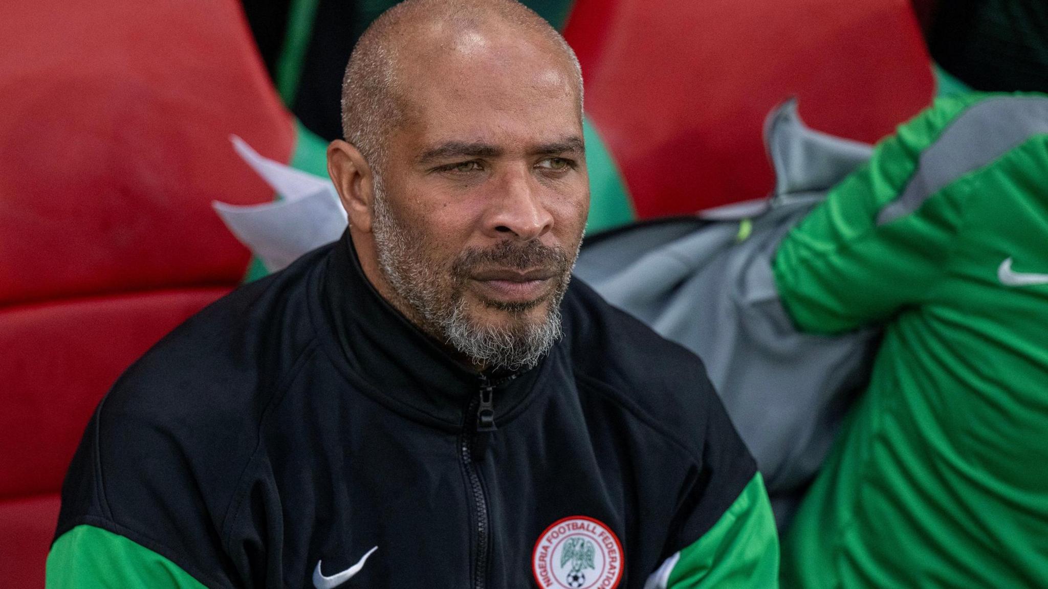 Eric Chelle is seen from the chest up wearing a black Nigeria tracksuit top with green sleeves and a federation crest on the chest. He sits on a red dugout chair while next to him a fellow coach can be seen putting a grey top on over a green t-shirt