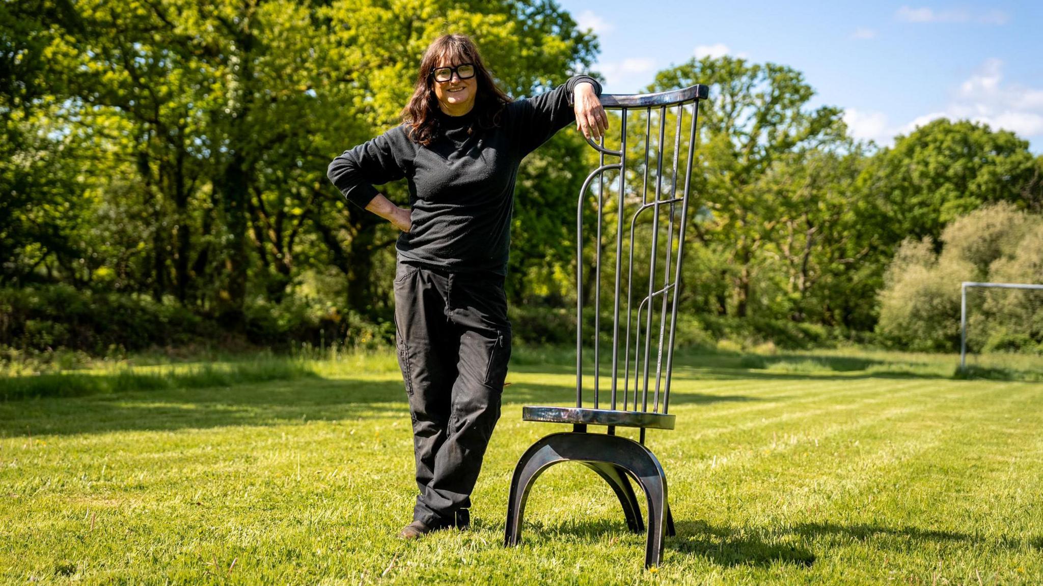 Woman in field leaning on a chair. She is wearing black trousers and a black long sleeve jumper. She has black glasses on.