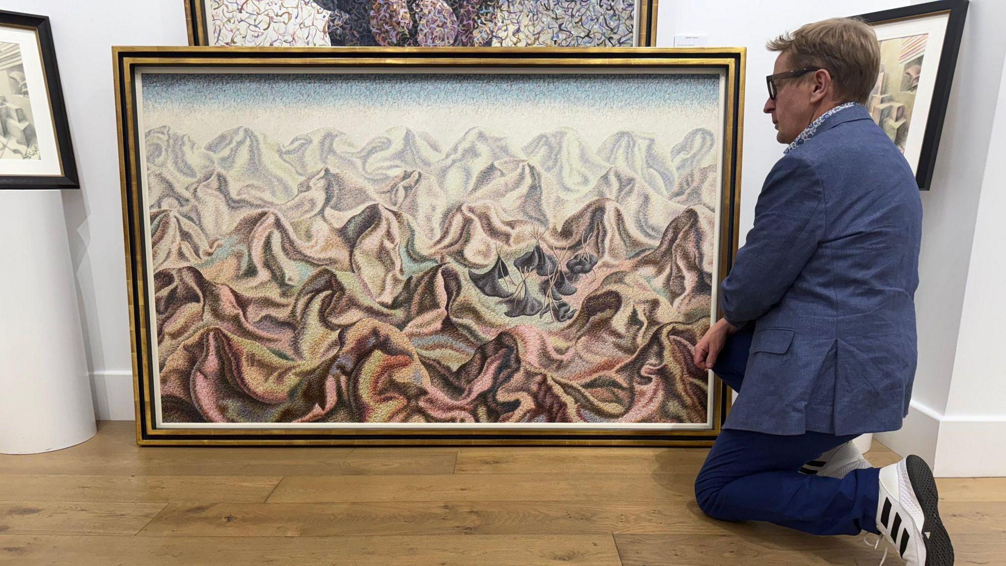 Art dealer Grant Ford bends down beside a surrealist Henry Orlik painting of a mountainous landscape 