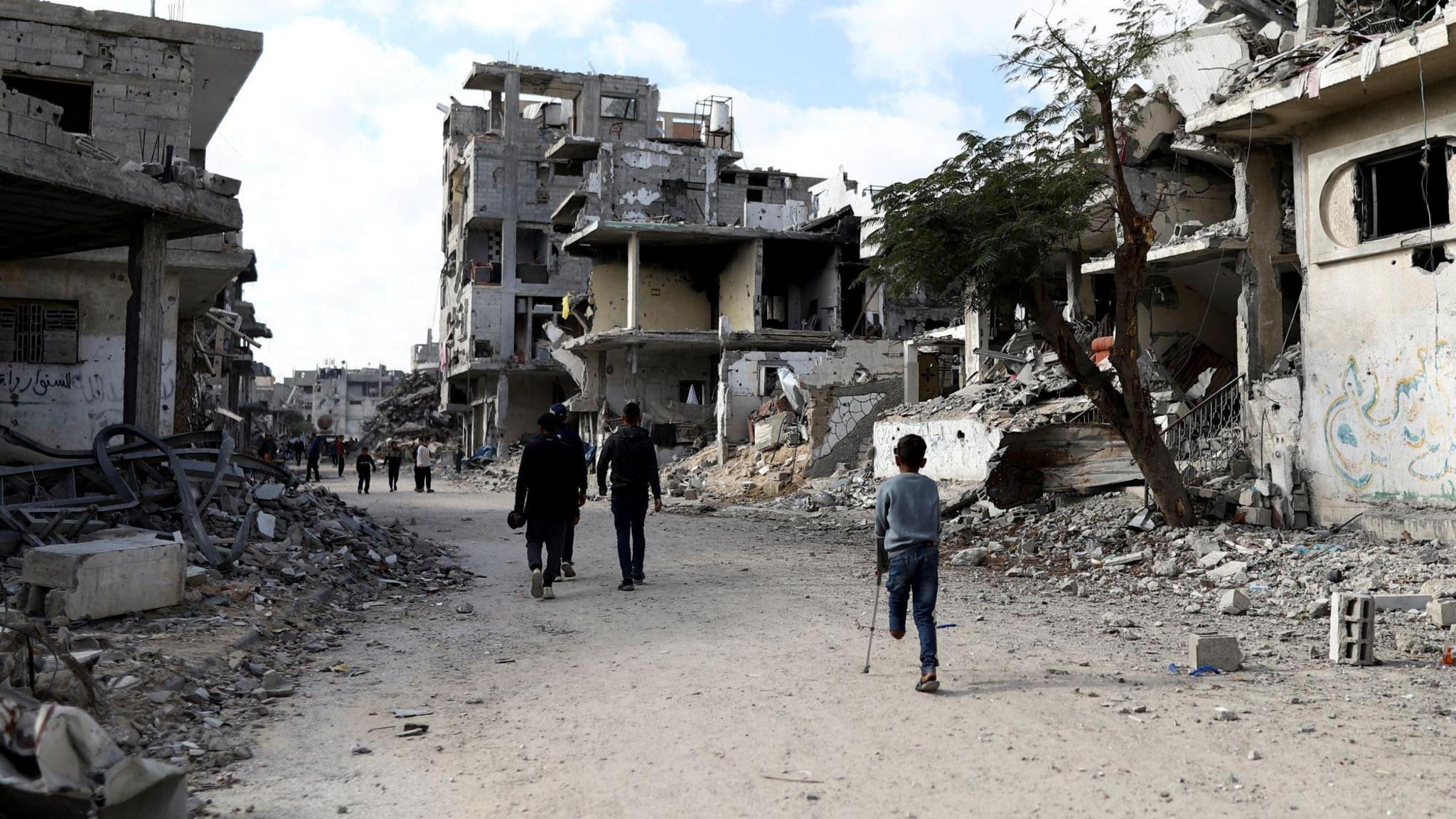 People, including a child on a crutch, missing the lower half of his left leg, walk past the grey, dusty rubble of houses and buildings destroyed during the war in Al-Bureij in central Gaza
