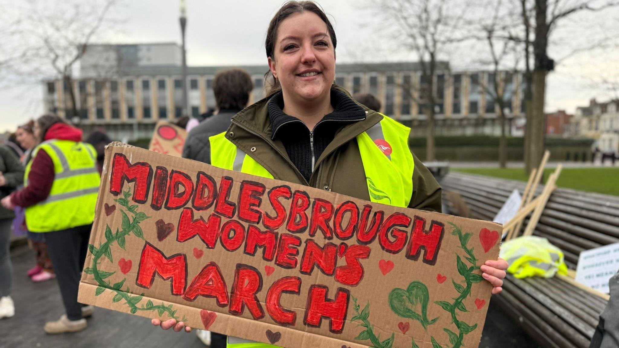 Lucy Wade smiling at the camera. She's got brown hair and is wearing a hi-vis vest. She's holding a placard that reads 'Middlesbrough Women's March'.