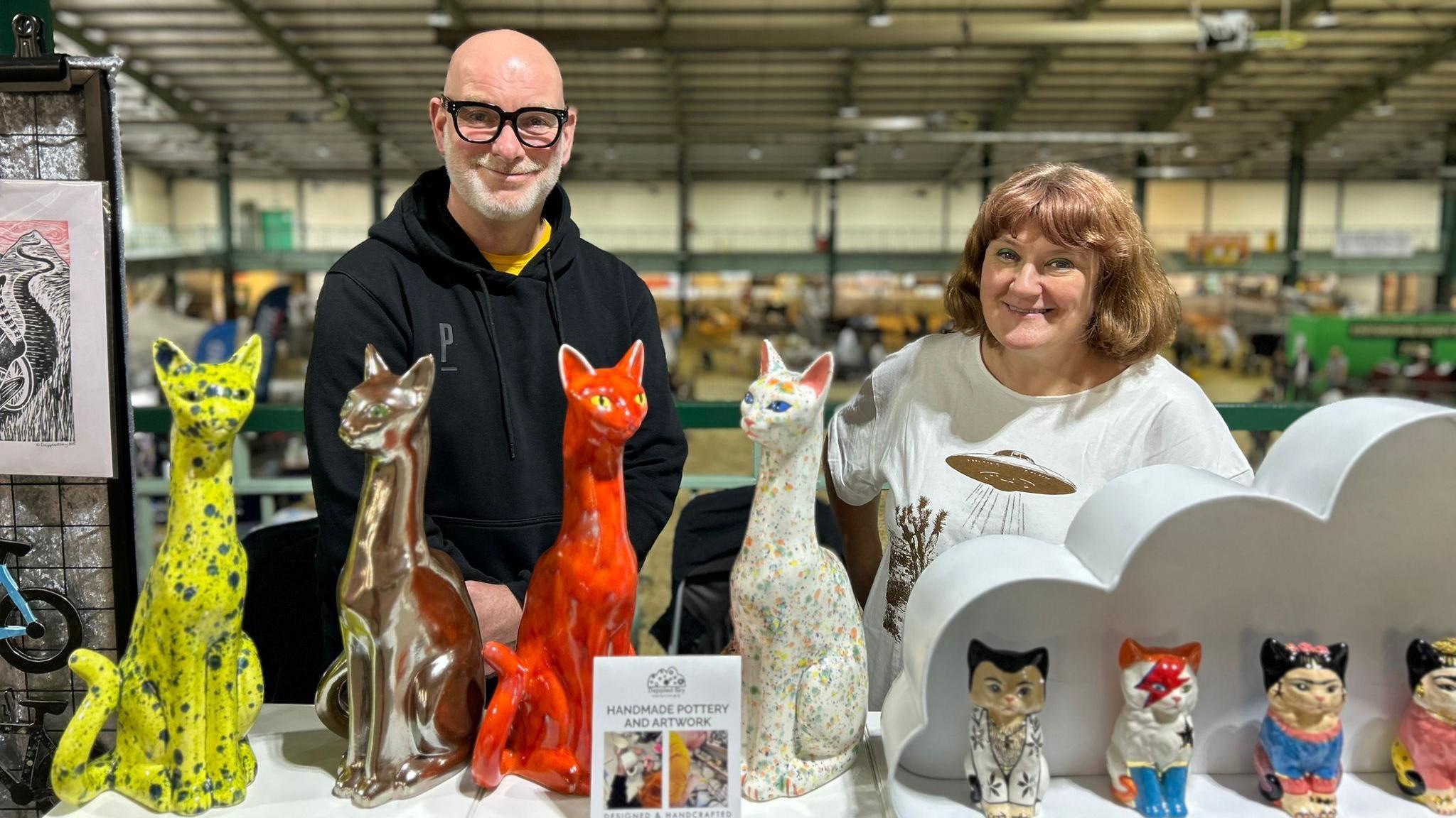 A man wearing glasses and a navy blue jacket is stood next to a woman wearing a white top. They are standing behind a range of pottery cats of various sizes.