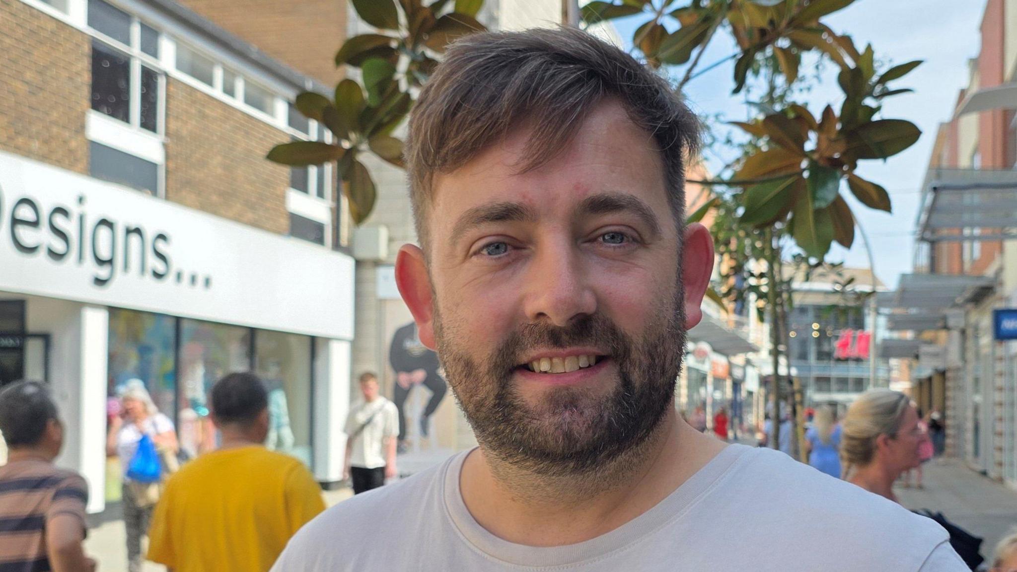 Jack Roper is standing in a busy shopping street. He has a white t-shirt on.