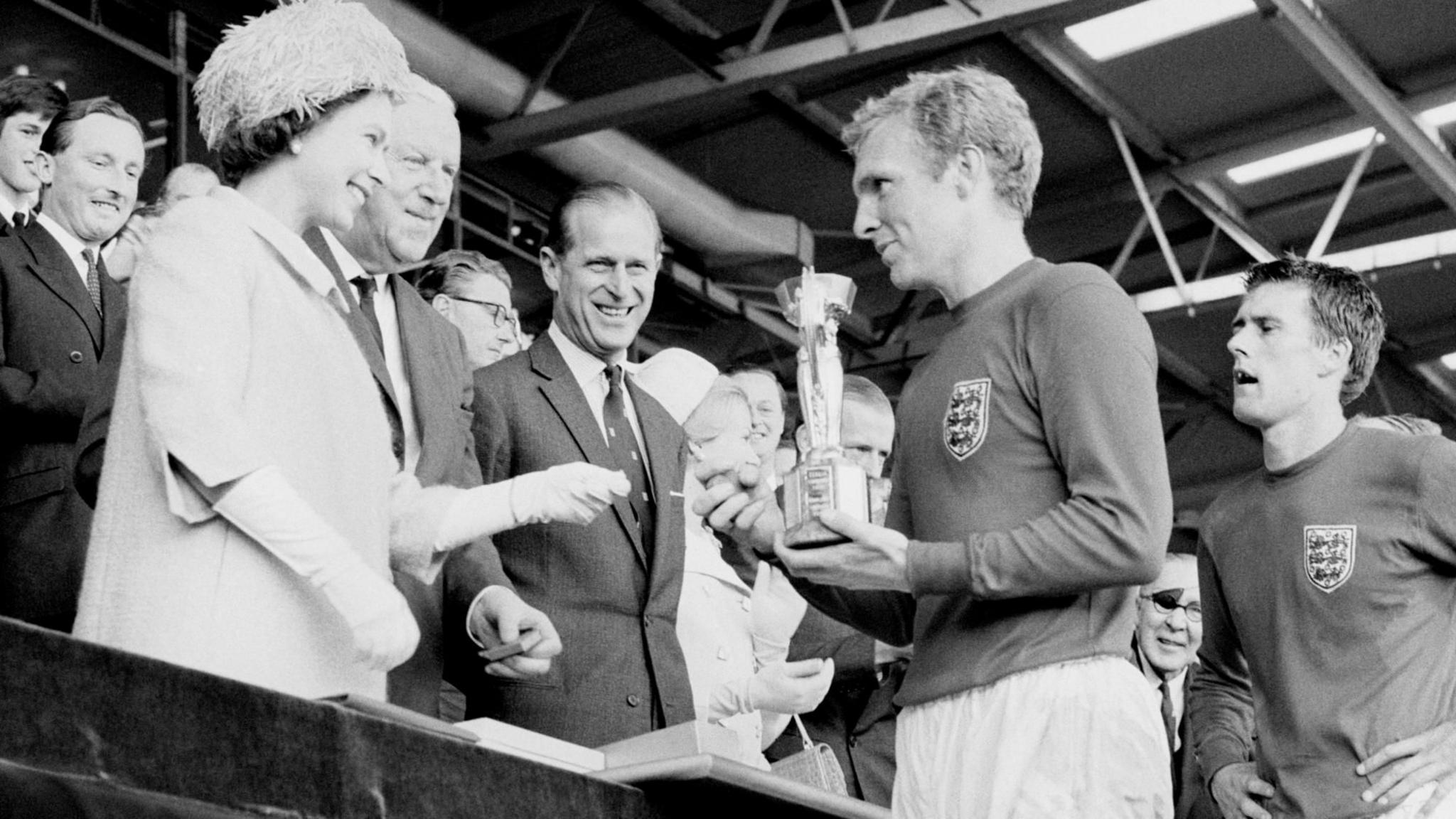 The Queen hands the World Cup trophy to England captain Bobby Moore in the Royal Box at Wembley Stadium. The black and white photo shows the late queen in a fluffy bonnet and smiling. Prince Philip also looks on smiling. Geoff Hurst stands behind Moore, waiting to collect his medal.
