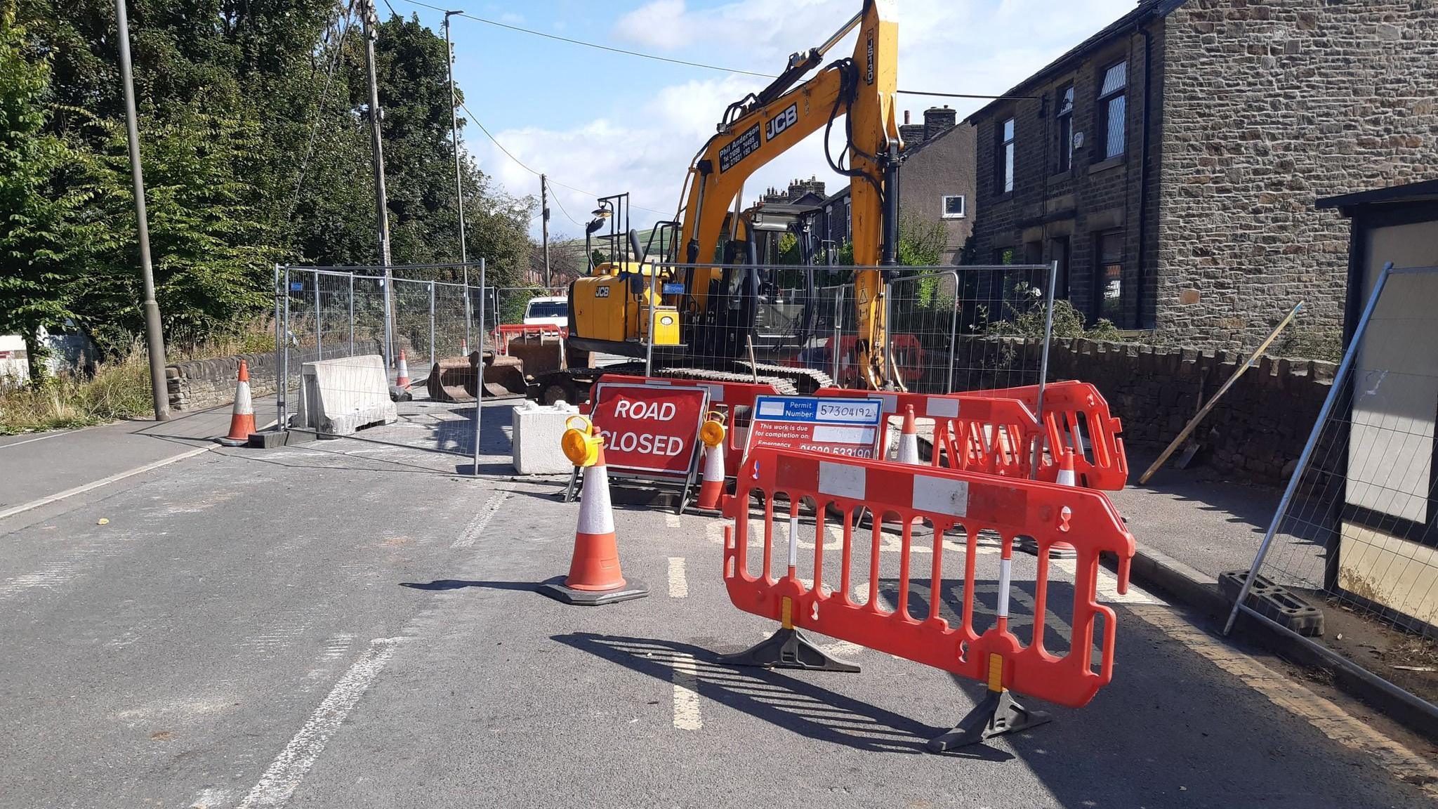 Fencing, concrete barriers, cones and a road cflosed sign show the blocked street, with a large yellow digger parked behind the fence. Housing is on the right hand side and trees on the left