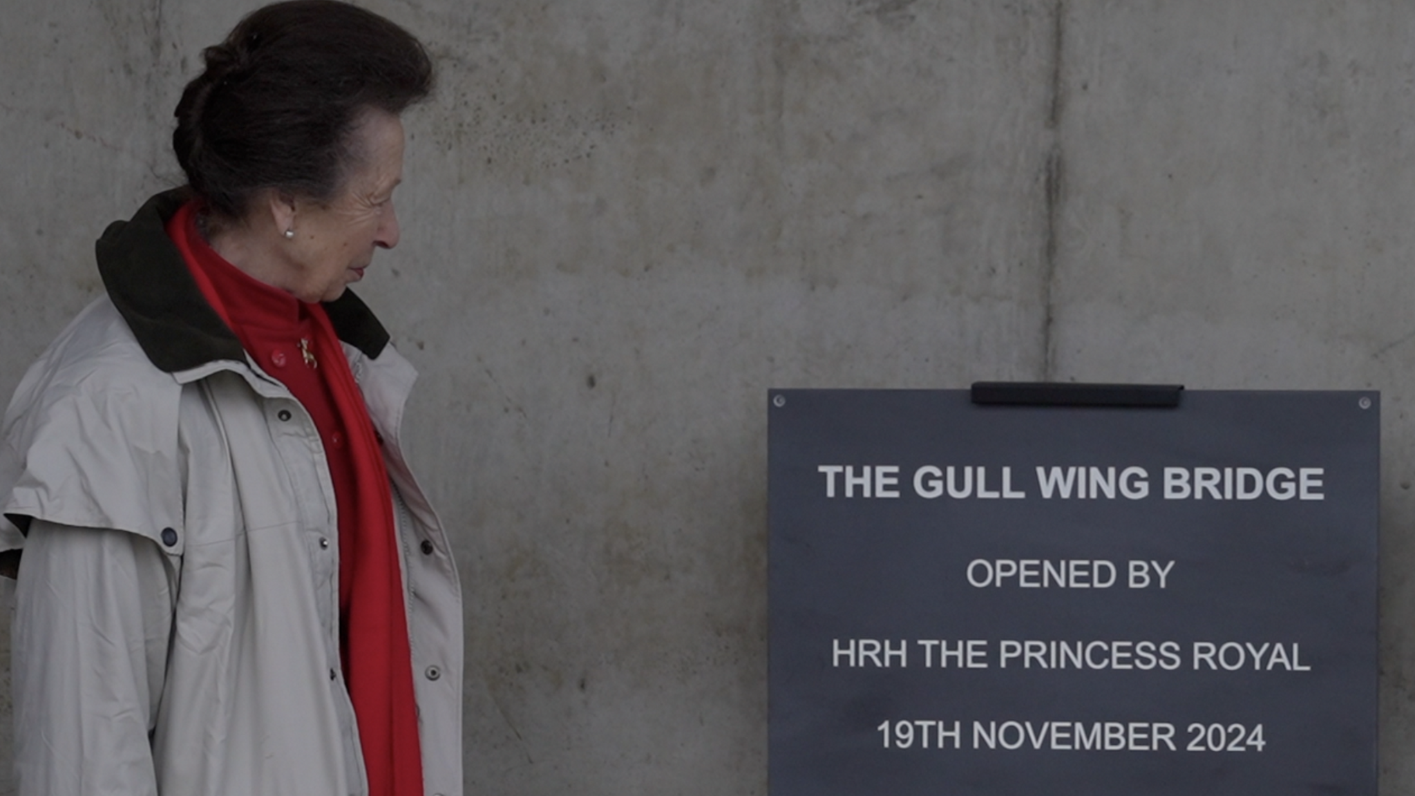 Princess Anne is pictured looking at a sign detailing the name of the bridge and the date she unveiled it. She has short dark hair and is wearing a red top with a red scarf and cream coat.