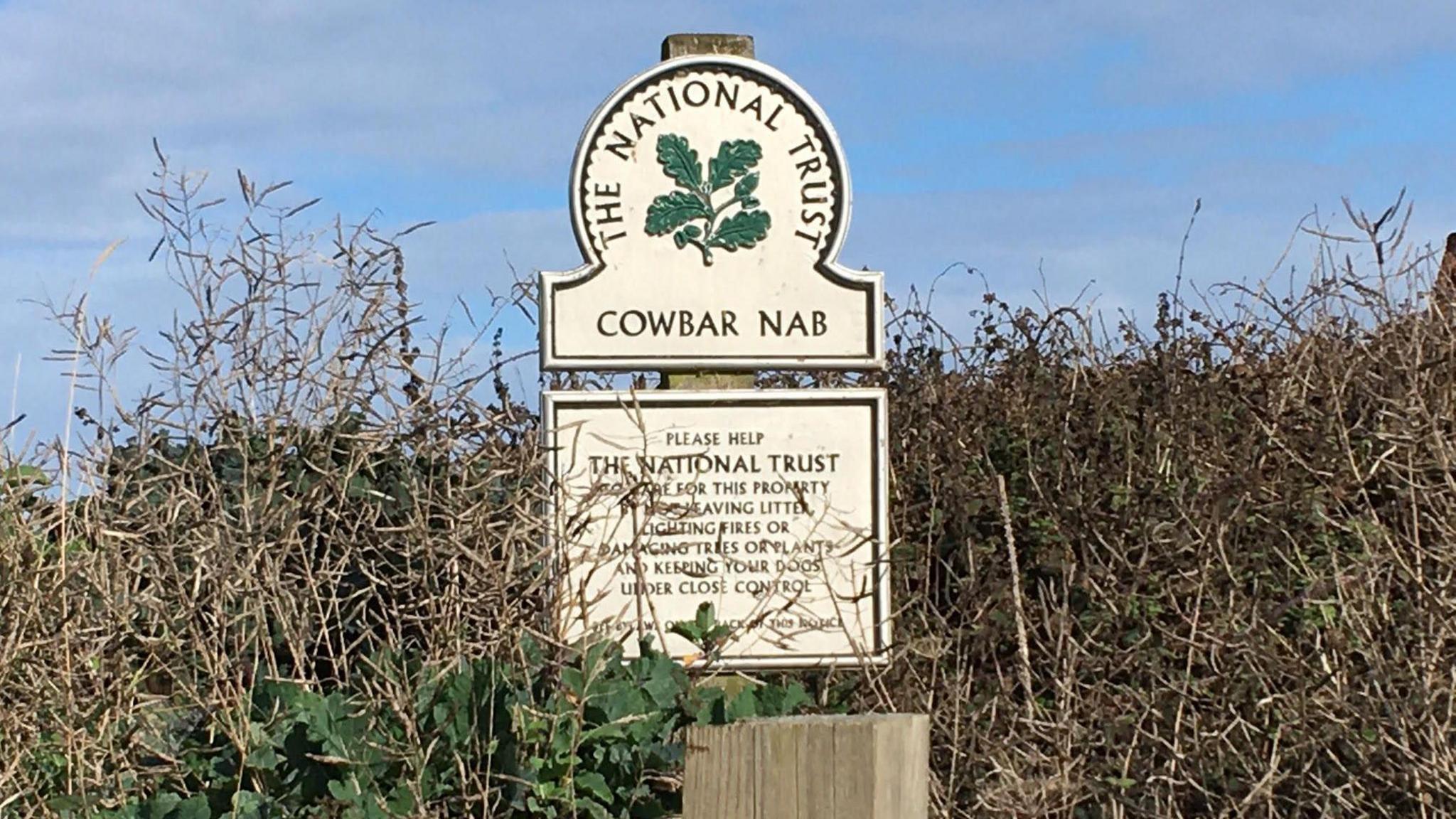 Cowbar, near Staithes, is adjacent to the Cleveland Way and a popular spot particularly with walkers. A National Trust sign stands in shrubbery with green lettering and oak leaves. The sign reads 'COWBAR NAB'.