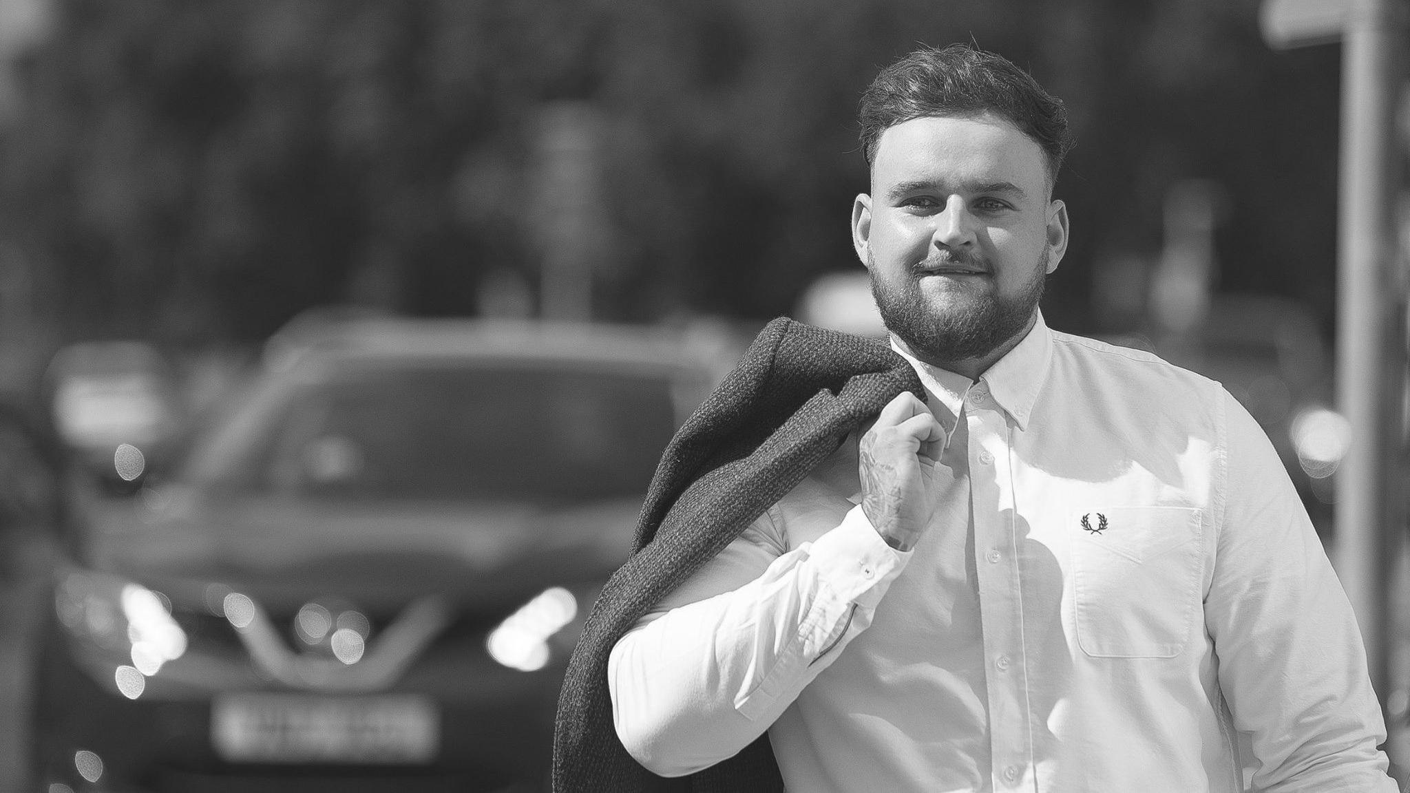 Luke Allen in a black and white photo. He is wearing a shirt and carrying a jacket over his shoulder.