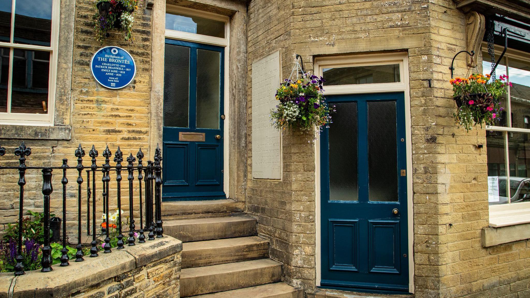 The front of a house, with two blue doors. A set of steps leads up to the door on the left. Next to it sits a blue plaque.