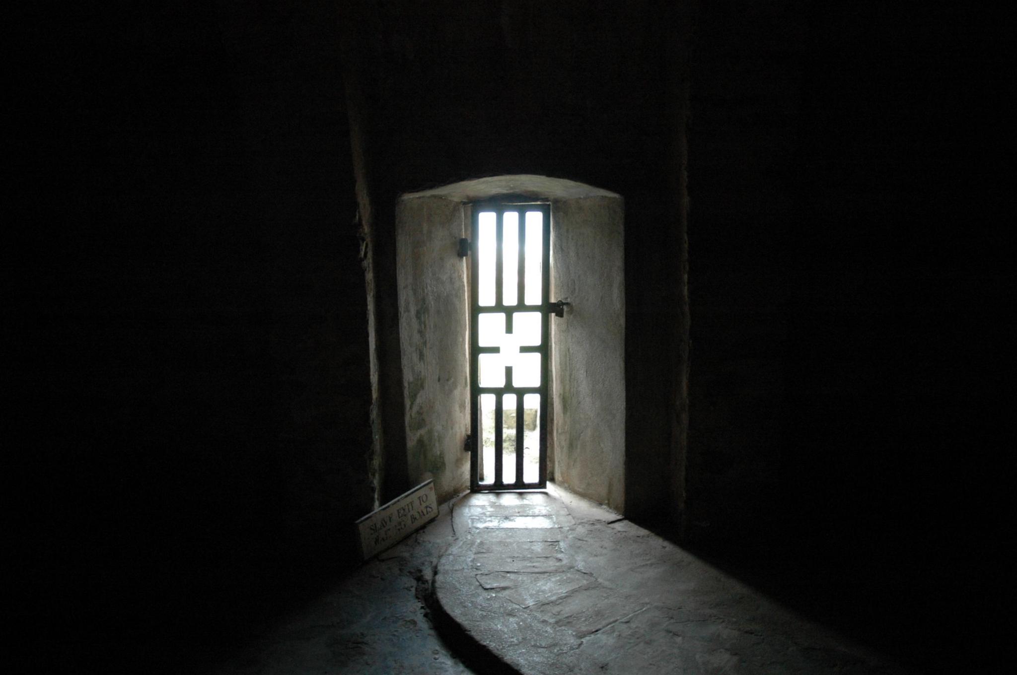 Sunlight is seen through a narrow door opening. The door is made up of metal bars. The walls above and to either side are dark.