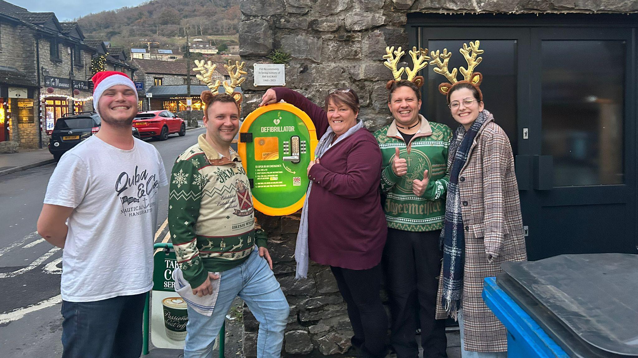 Tina Salway stands with three male volunteers in front of a green and yellow defibrillator that's been installed on a stone wall outside a business. 