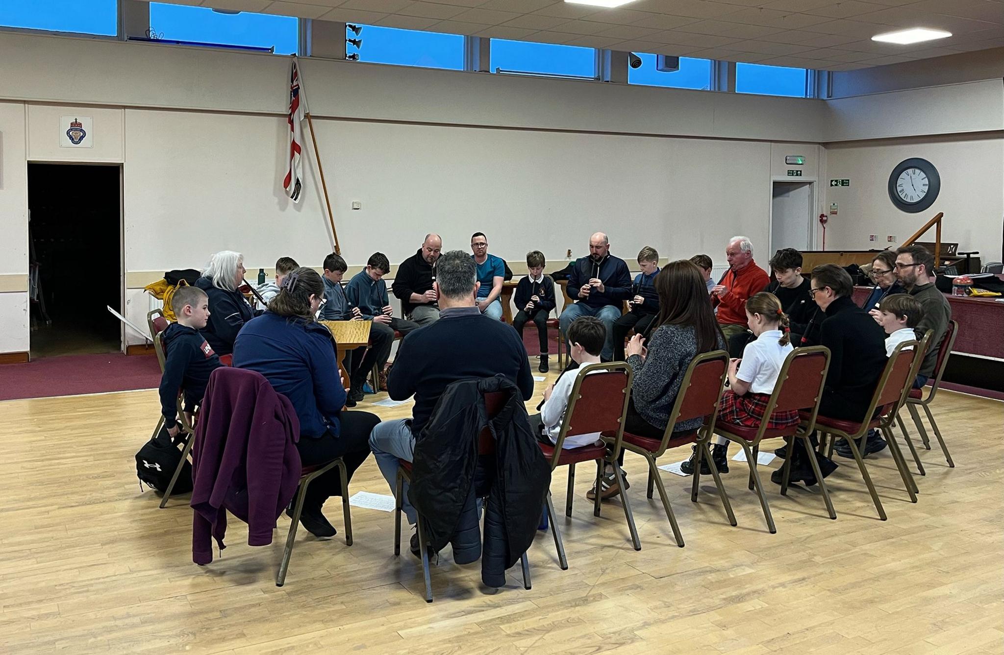 A group of people learning to play the bagpipes using changers sit on chairs in a circle in a large hall. 