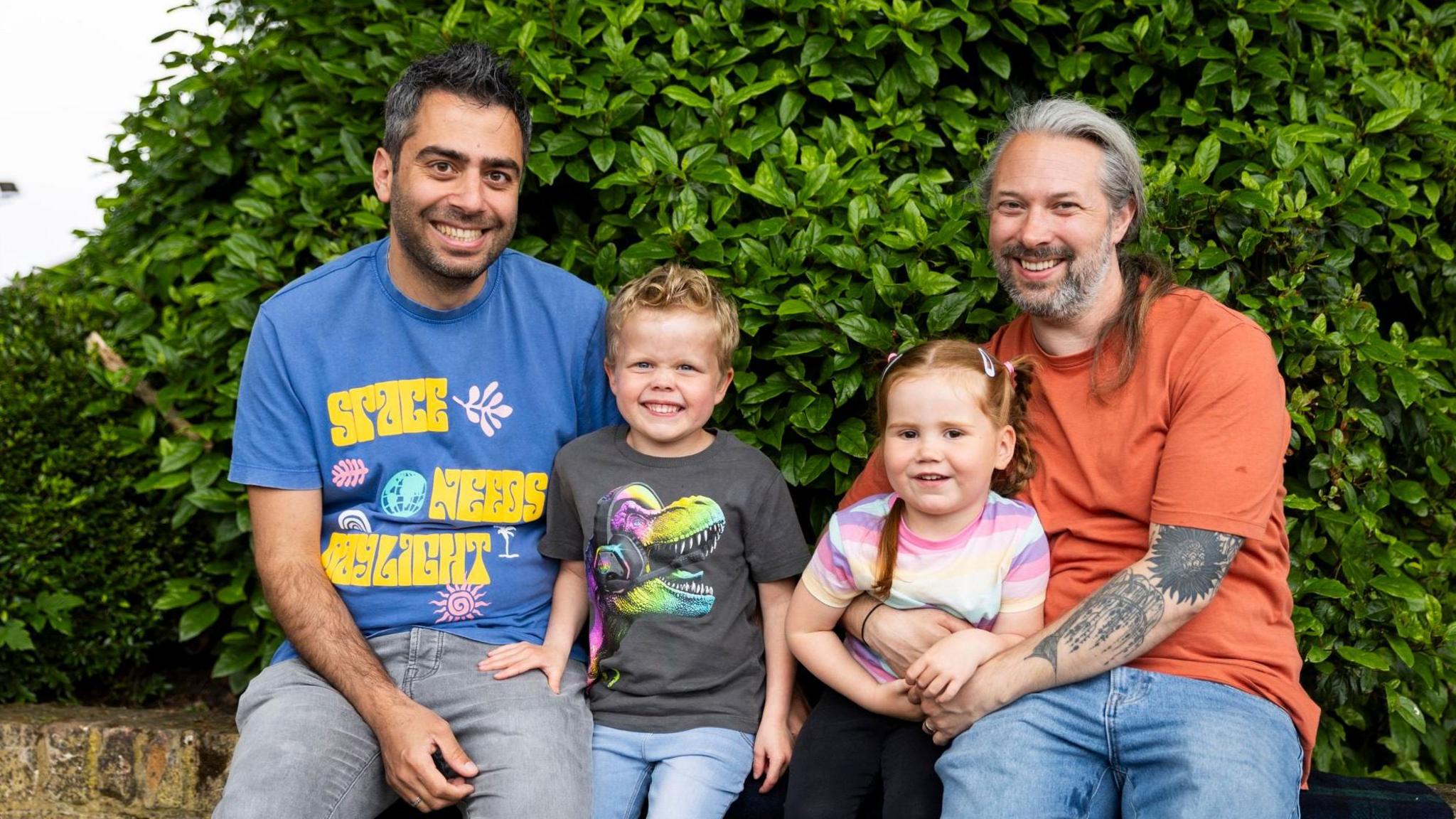 Luke and Alex sitting on a brick wall with a hedge behind them. In the middle are their two young daughters. They are all smiling at the camera.