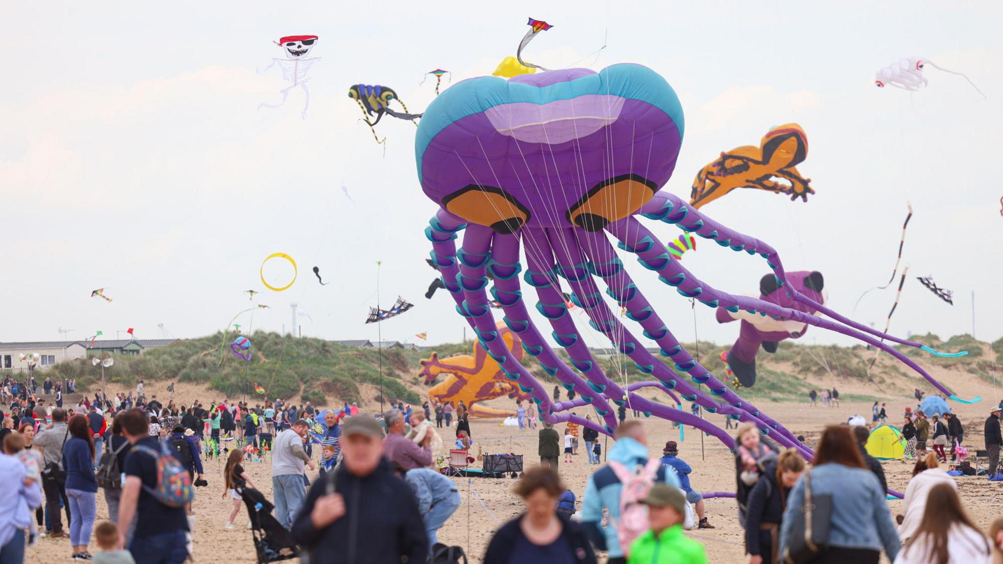 A large kite in the shape of an octopus being flown on Redcar beach. It is purple with large orange eyes. A number of smaller kites are also being flown in the area with dozens of people looking on.