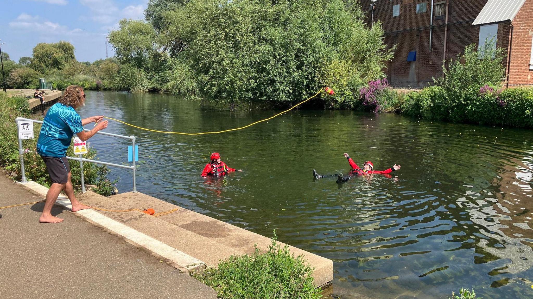 A man is throwing a floatation ring toward two people dressed in red floating in a river