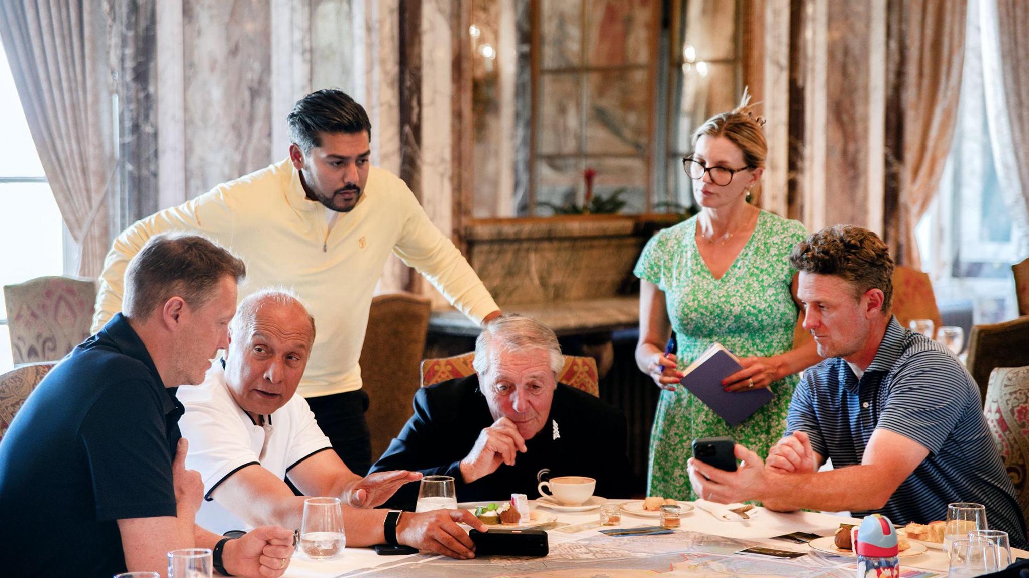 Four men sitting around a table with one man standing behind and leaning over and a woman standing and holding a large notepad. There are glasses and cups on the table.