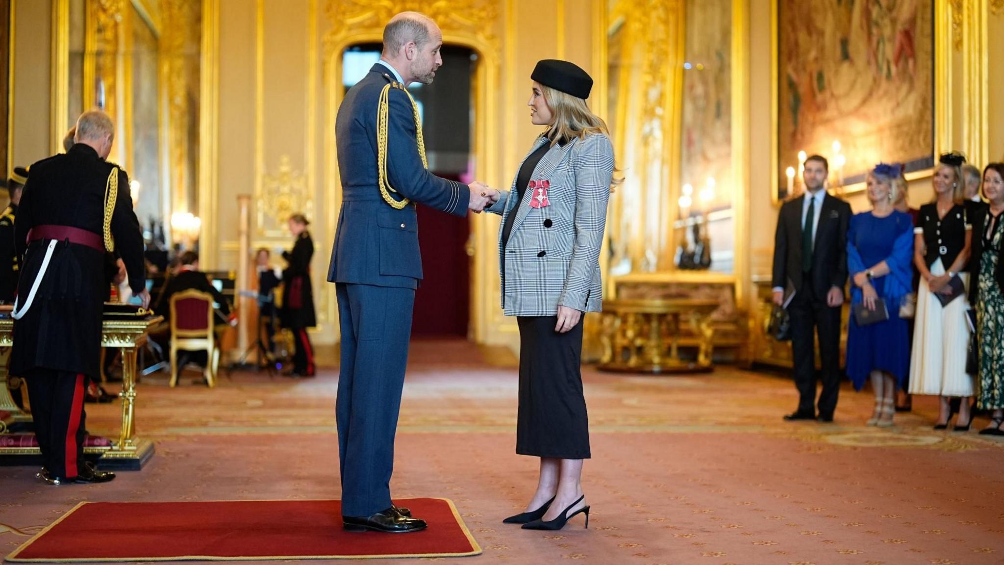 Georgia Harrison shaking hands with Prince William as she is presented with her MBE medal, which is pinned to her jacket breast pocket. The prince is standing on a square of red carpet and is wearing a blue uniform with circular lengths of gold braid hanging on his right shoulder and going under his armpit. Harrison is standing off the carpet in front of him. They are in a grand room which has a gold glow. Other people are standing in line on the right and appear to be waiting to be seen next, and there are castle staff behind the prince and in the background