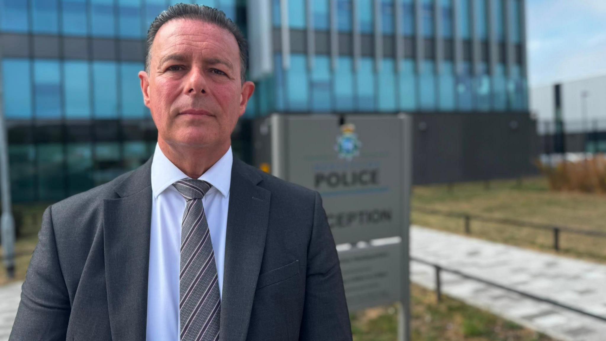 A middle aged man with short, greying black hair, in a dark grey suit, white shirt and grey striped tie looks into the camera with a serious expression. He is standing in front of a paved pathway leading to a large office building, and there is a sign featuring the crest of Merseyside Police immediately behind him.