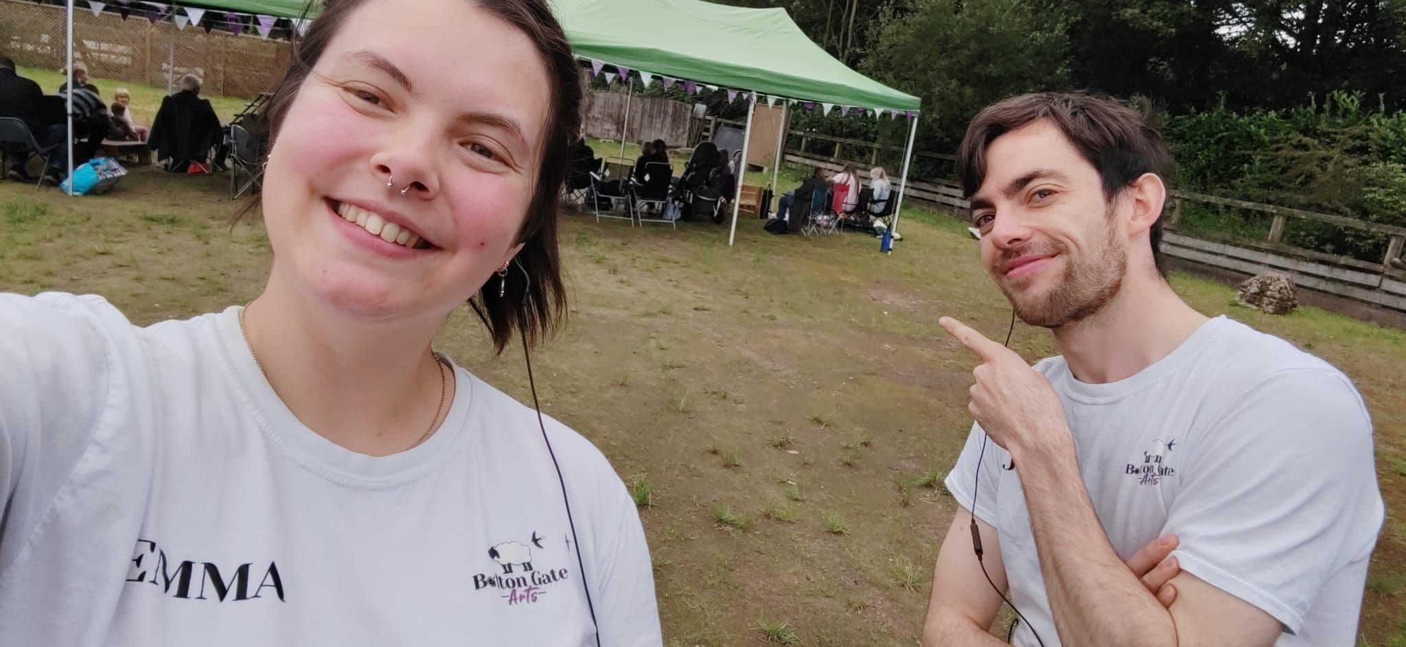 A selfie of two people stood in a field with a gazebo in the background. Beneath the gazebo, people sit in foldable chairs. The two people in the foreground are a man and woman wearing matching T-shirts with the logo of a farm. The woman has a nose ring and her name EMMA is also printed on her T-shirt.