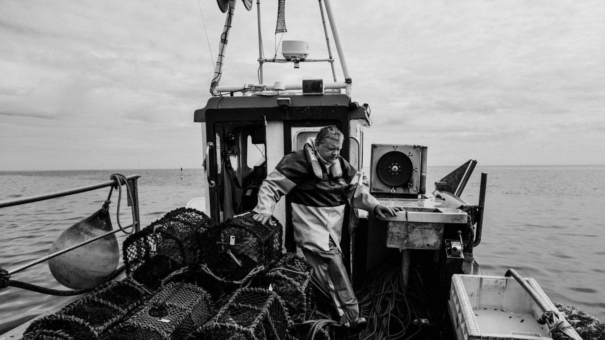 Black and white picture of a fisherman emerging from the wheelhouse of his small boat out at sea. He is walking while steadying himself on a large pile of lobster pots stacked on one side of the boat.