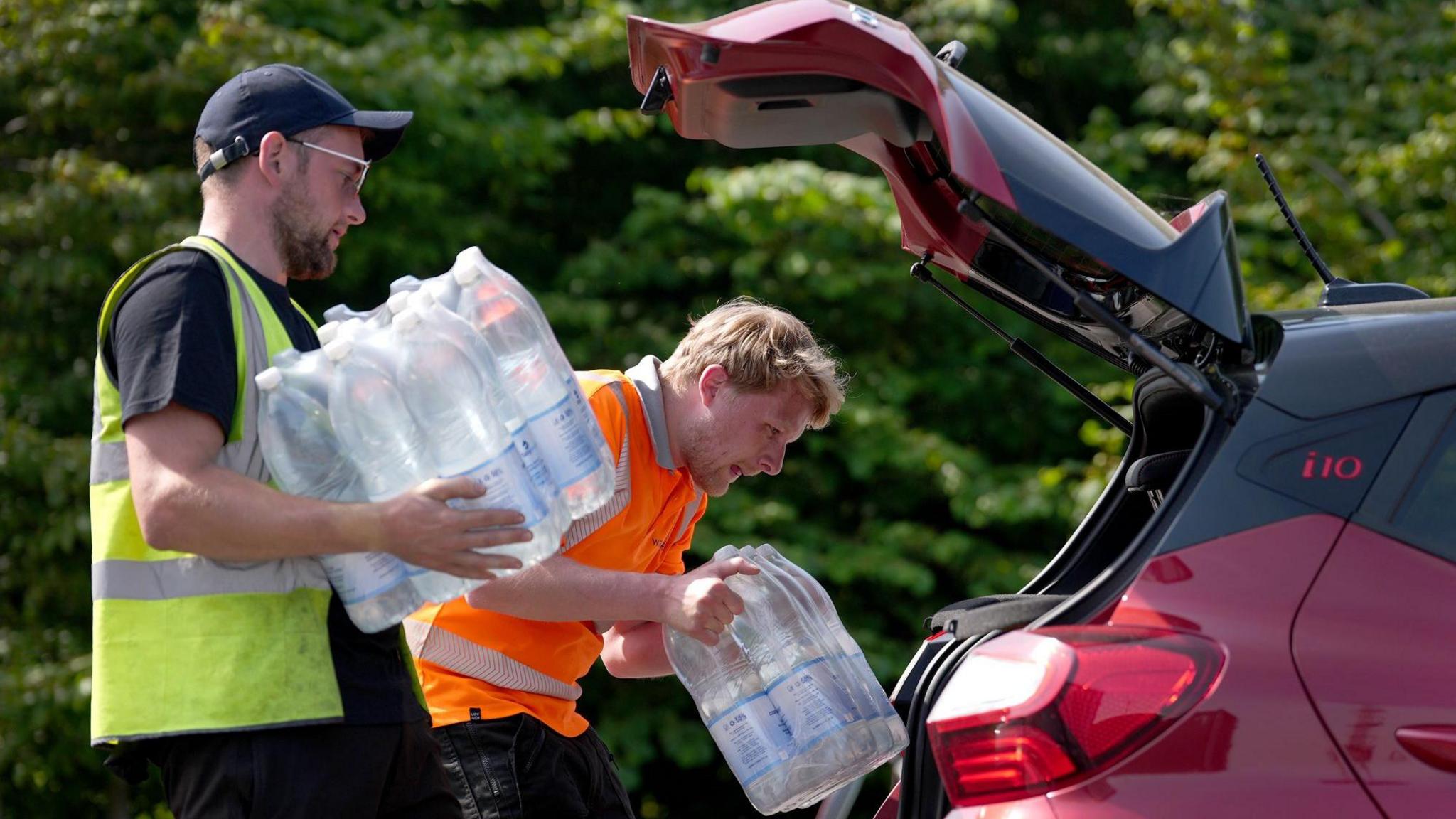 A man wearing a yellow his vis jacket and a man wearing an orange hi-vis jacket, each carrying a pallet of water, are in the process of putting it into the back of a red car.