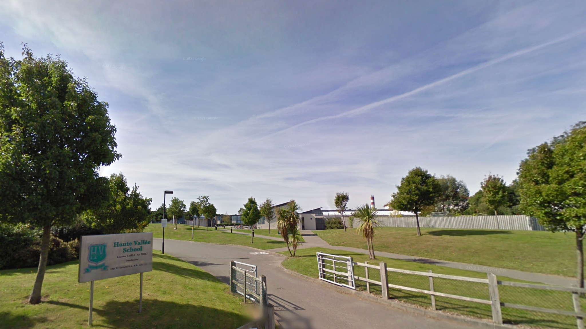 A Google image photo of the grasses and entry of Haute Vallée School. The Haute Vallée School sign is stuck in the grass to the left of the entrance with the school building in vision in the distance.