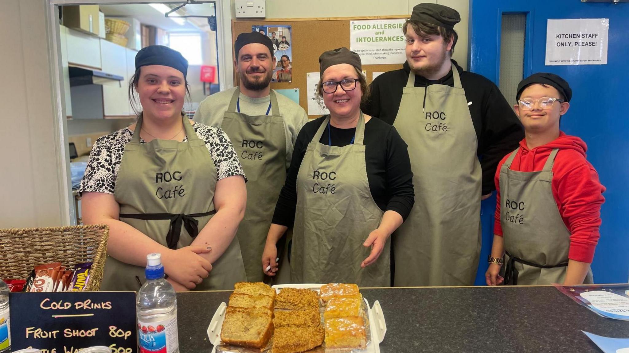 Five adults in aprons and chefs hats smile from behind a counter top at a cafe