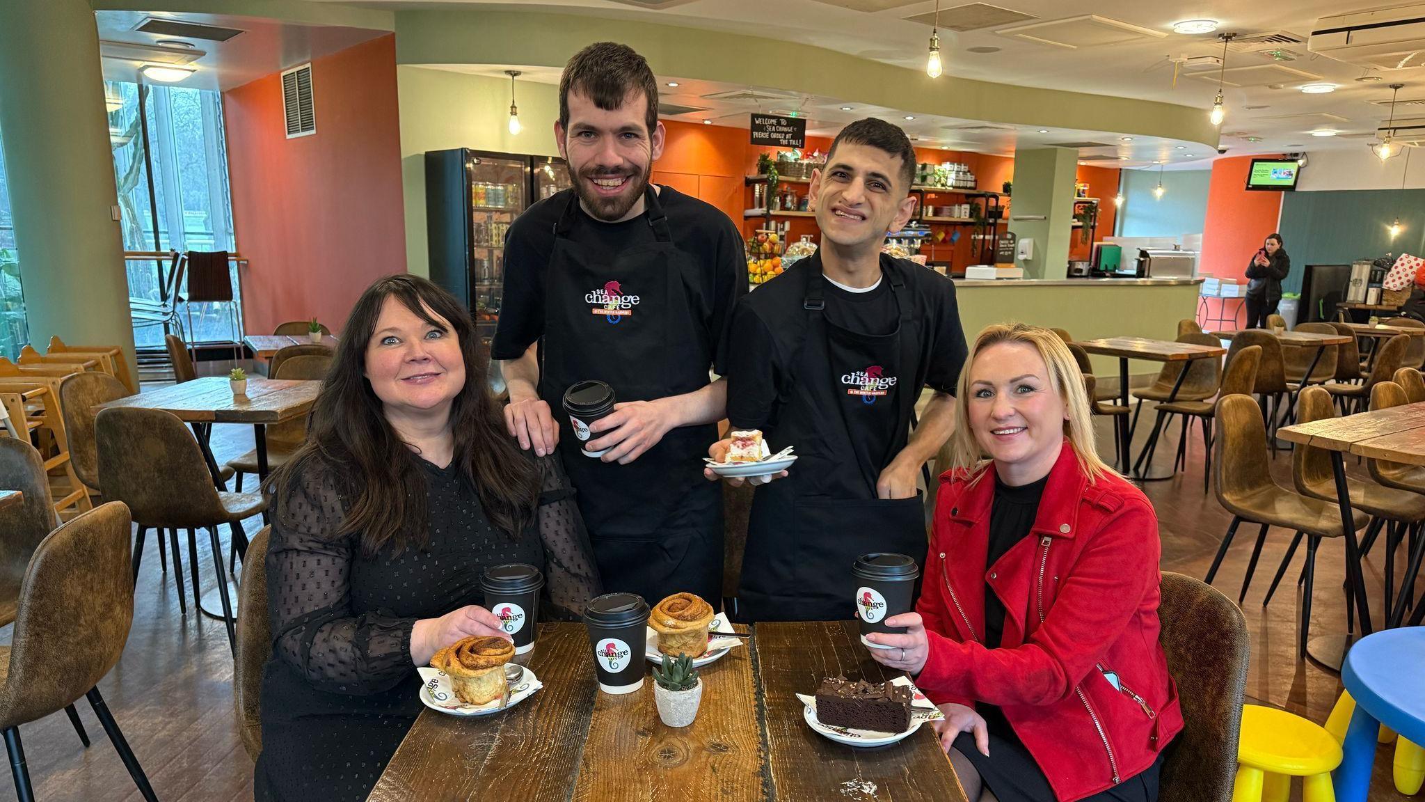 Sarah Farrell-Forster (left) is sitting at a wooden table with Councillor Beth Jones. They both have a selection of cakes and a hot drink and are smiling as they look into the camera. They are with two of the cafe workers, who also have items of food and drink and are smiling towards the camera.