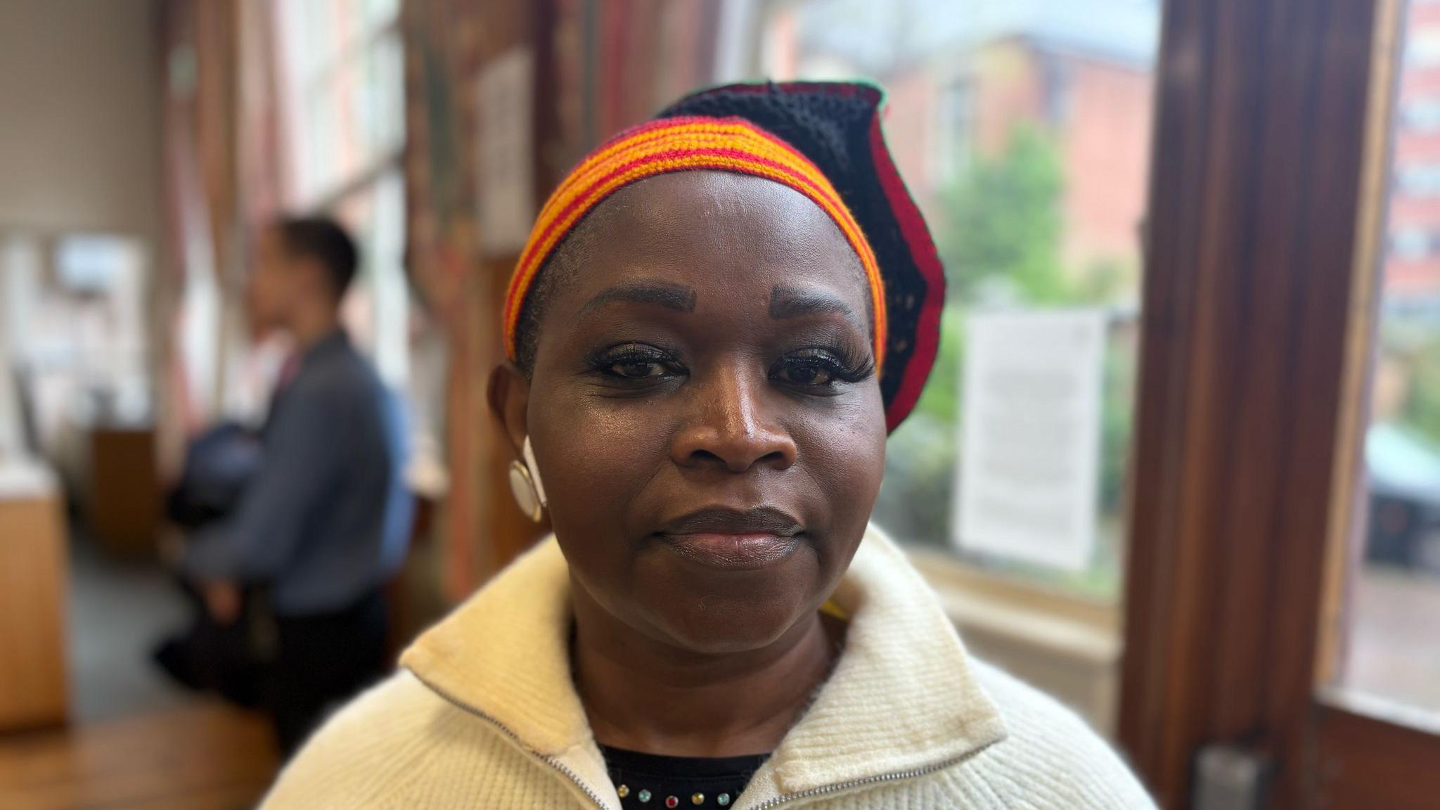 A close-up image of a woman looking at the camera. She wears a white jumper and a multi-coloured garment on her head.