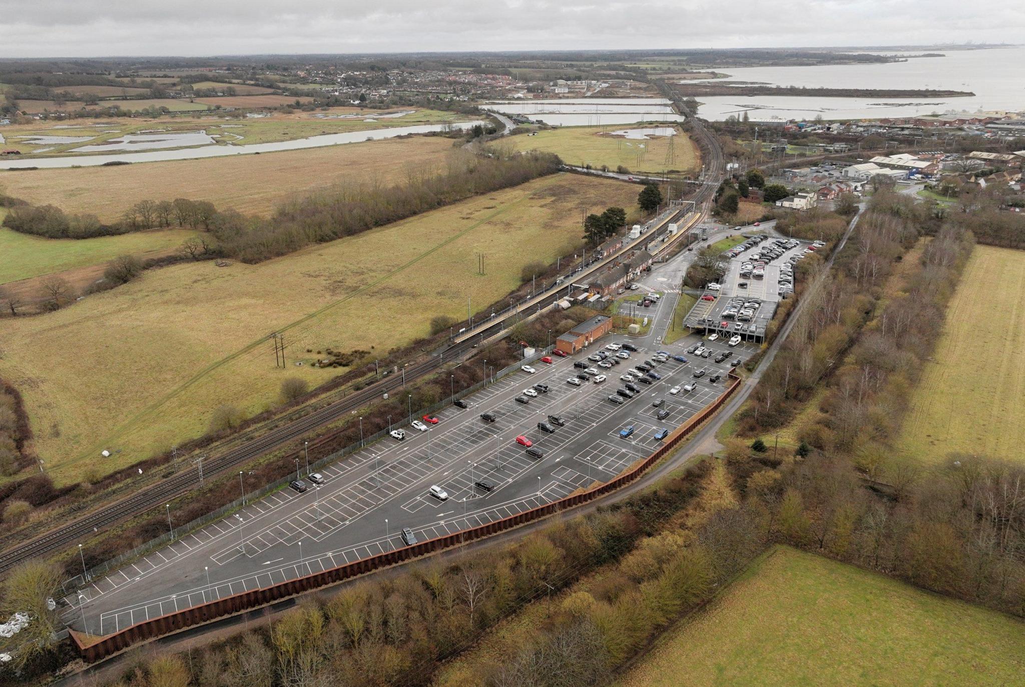 Aerial view of Manningtree station, showing large car park running alongside the railway line. A river and estuary can be seen behind it, along with fields and other buildings.