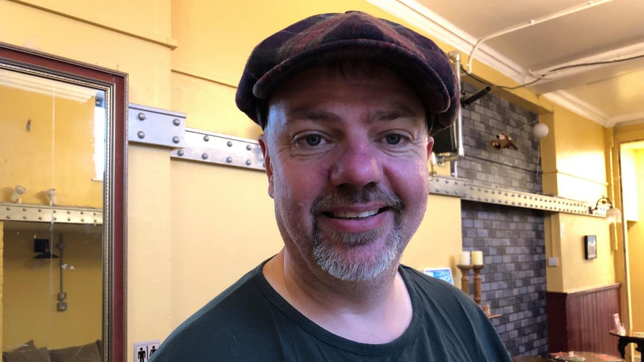 A man with a goatee beard and a flat cap is smiling at the camera. He is wearing a black T-shirt and is standing indoors in a cafe with cream-coloured walls. 