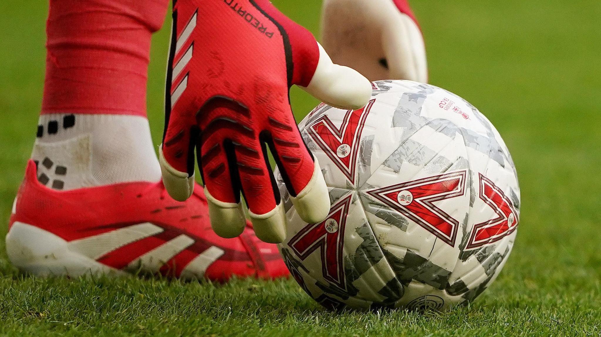 A close up of a football on grass. A goalkeeper wearing gloves leans down to pick the ball up.