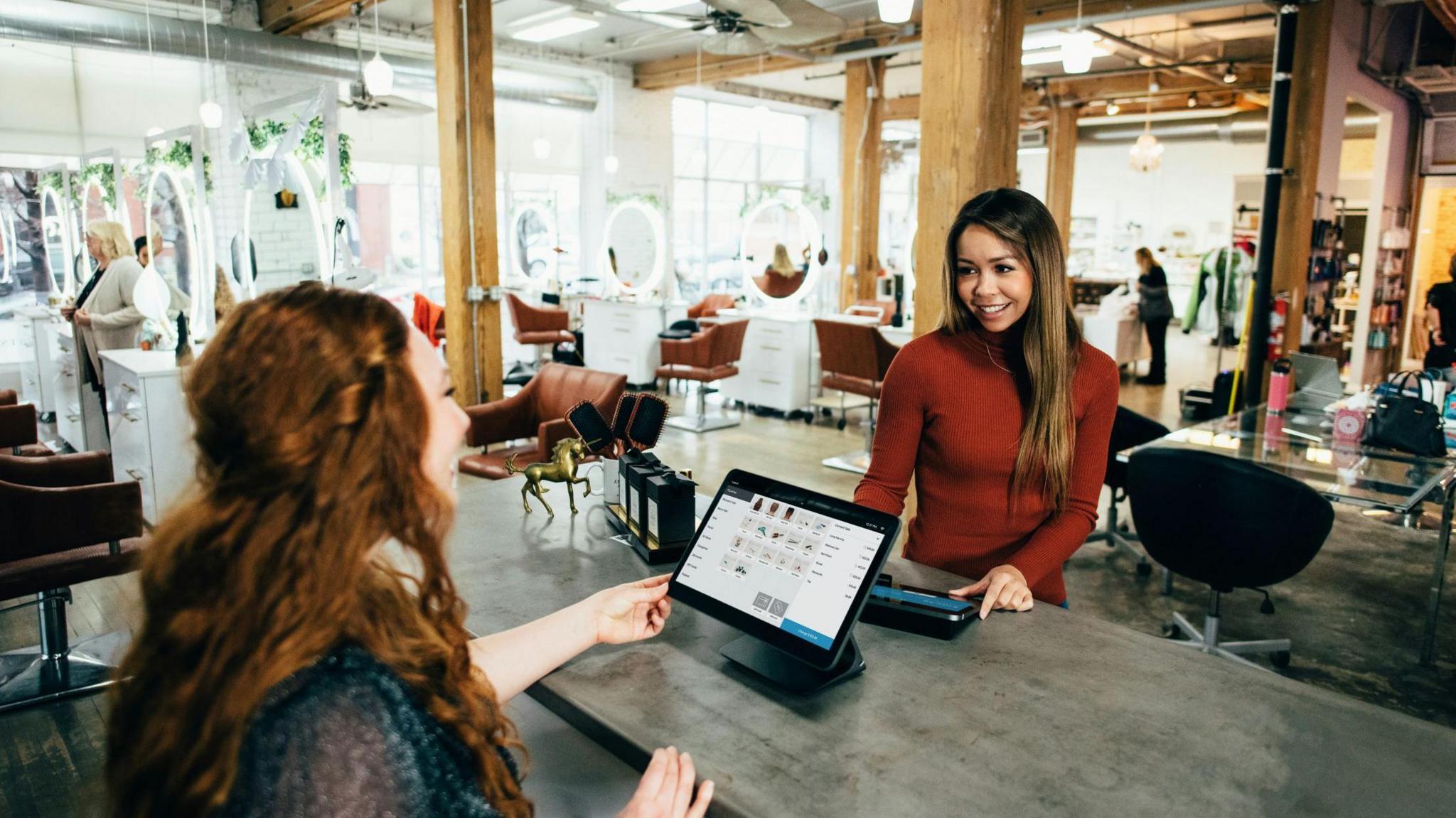 A stock image showing the interior of a bright, modern hair salon. In the foreground, two women are interacting at a counter — one stands behind it, smiling, while the other faces her from the customer side, holding what looks like a payment terminal. The counter has a computer screen and some small items on display. Behind them, the salon is open and well-lit with large mirrors, chairs, and people working or chatting. The space features wooden beams, exposed ceilings, and large windows letting in natural light.