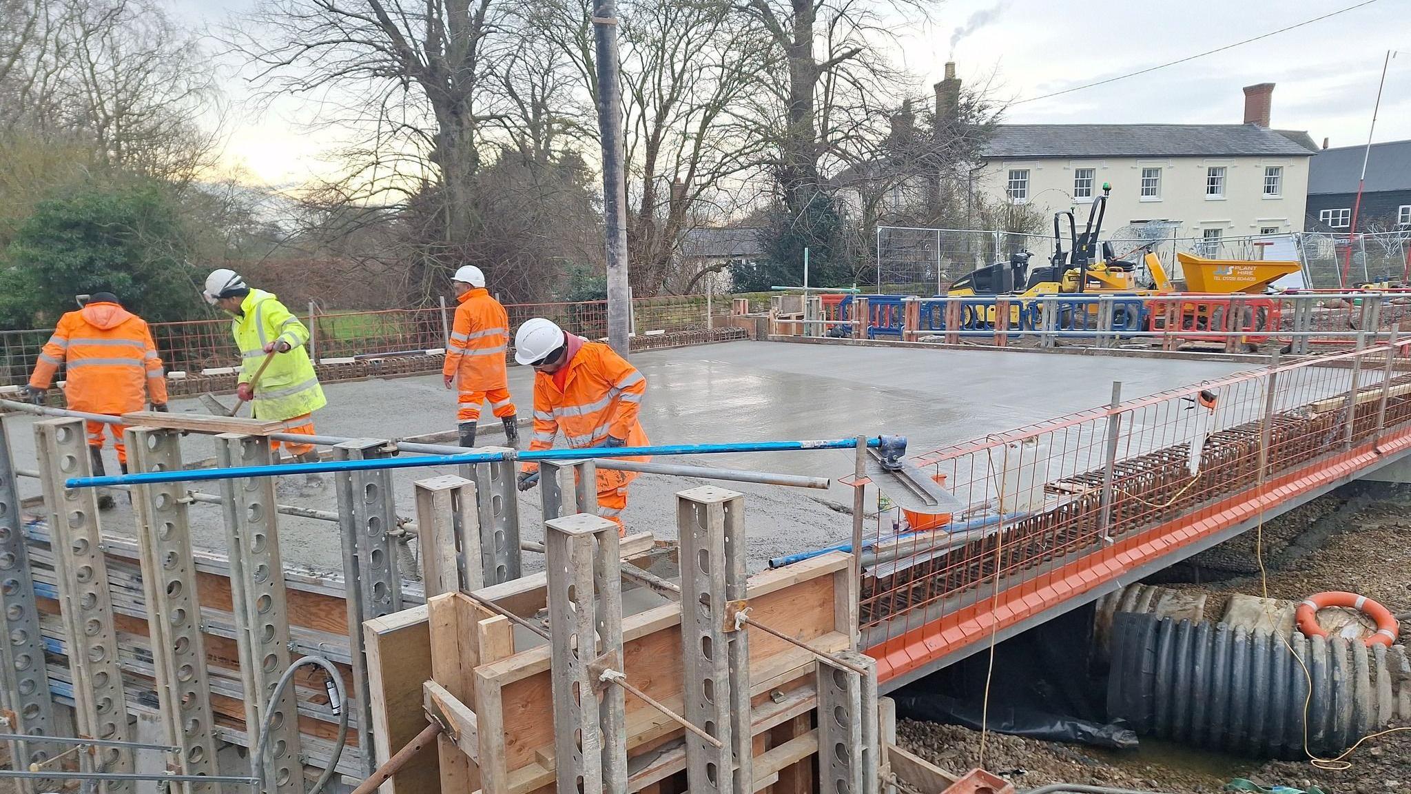 Workers wearing orange and yellow hi-vis coats and trousers and white hard hats working on the new bridge. The area has been cordoned off and machinery is in the distance.