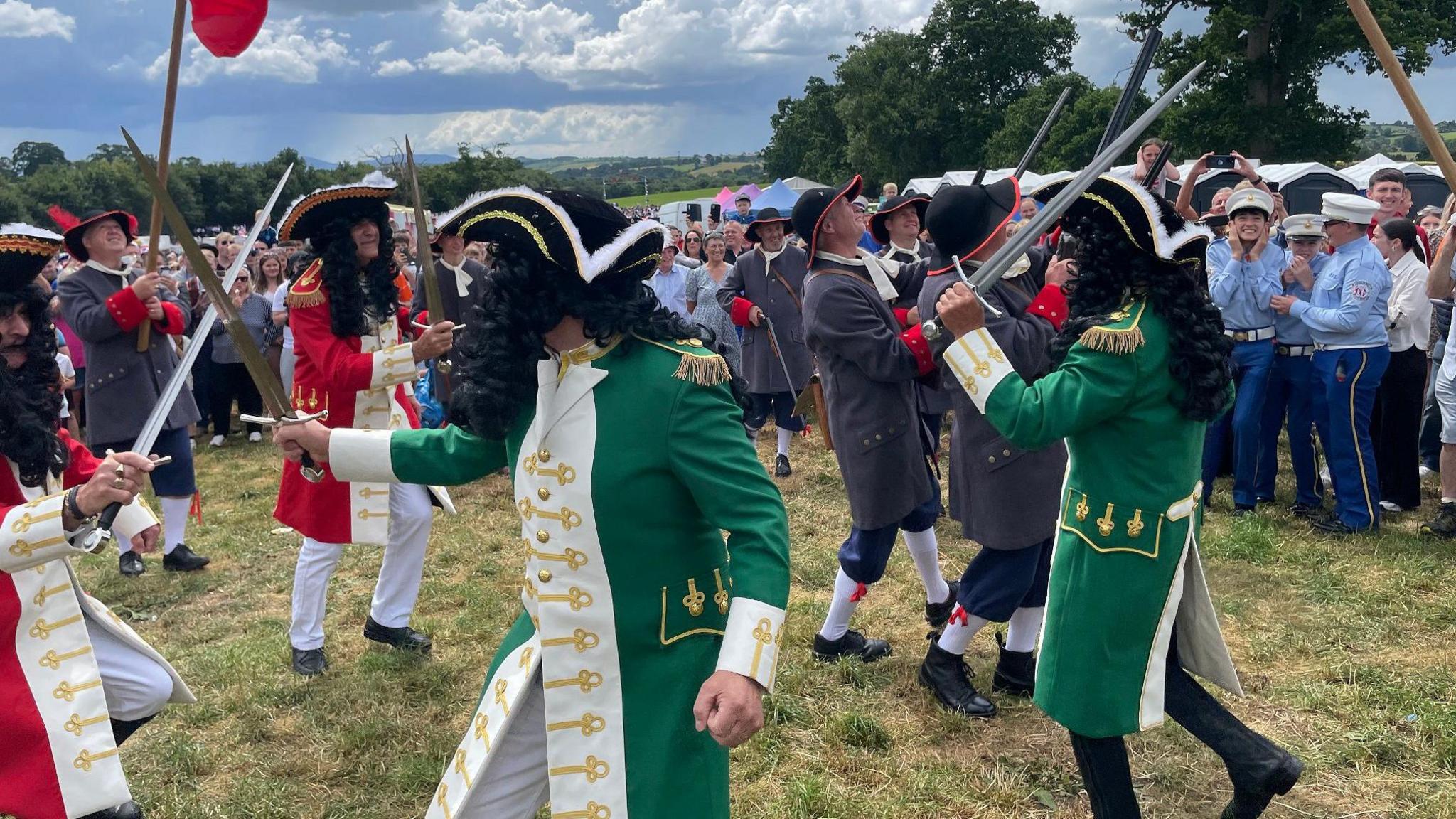 A group of men dressed as 17th century soldiers re-enact a the Battle of the Boyne. Some dressed in wither red and white or green and white uniforms are armed with swords, while some dressed in grey are armed with muskets. 