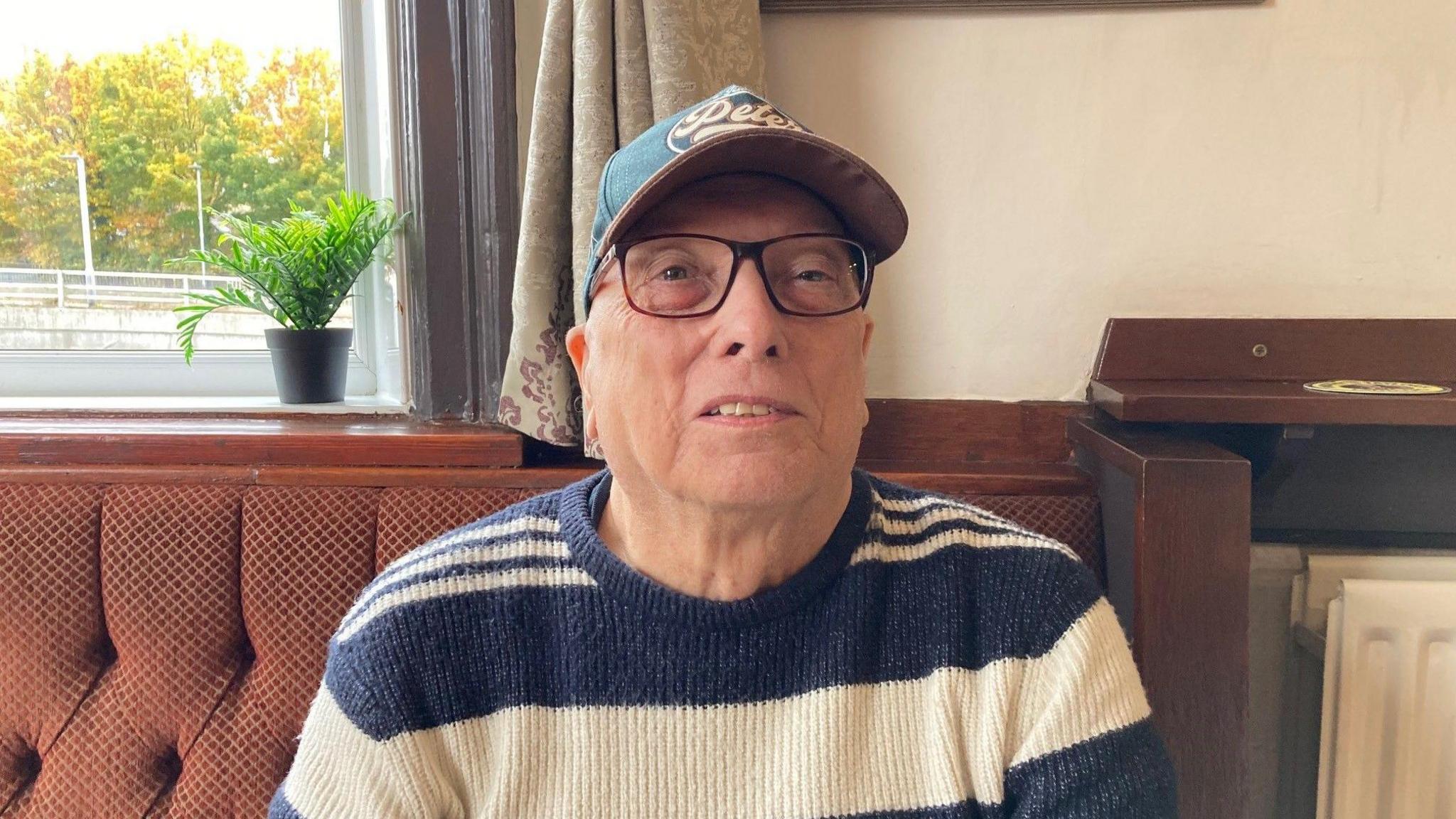 A man wearing a baseball cap. glasses and a blue and white striped jumper on some banquette seating.