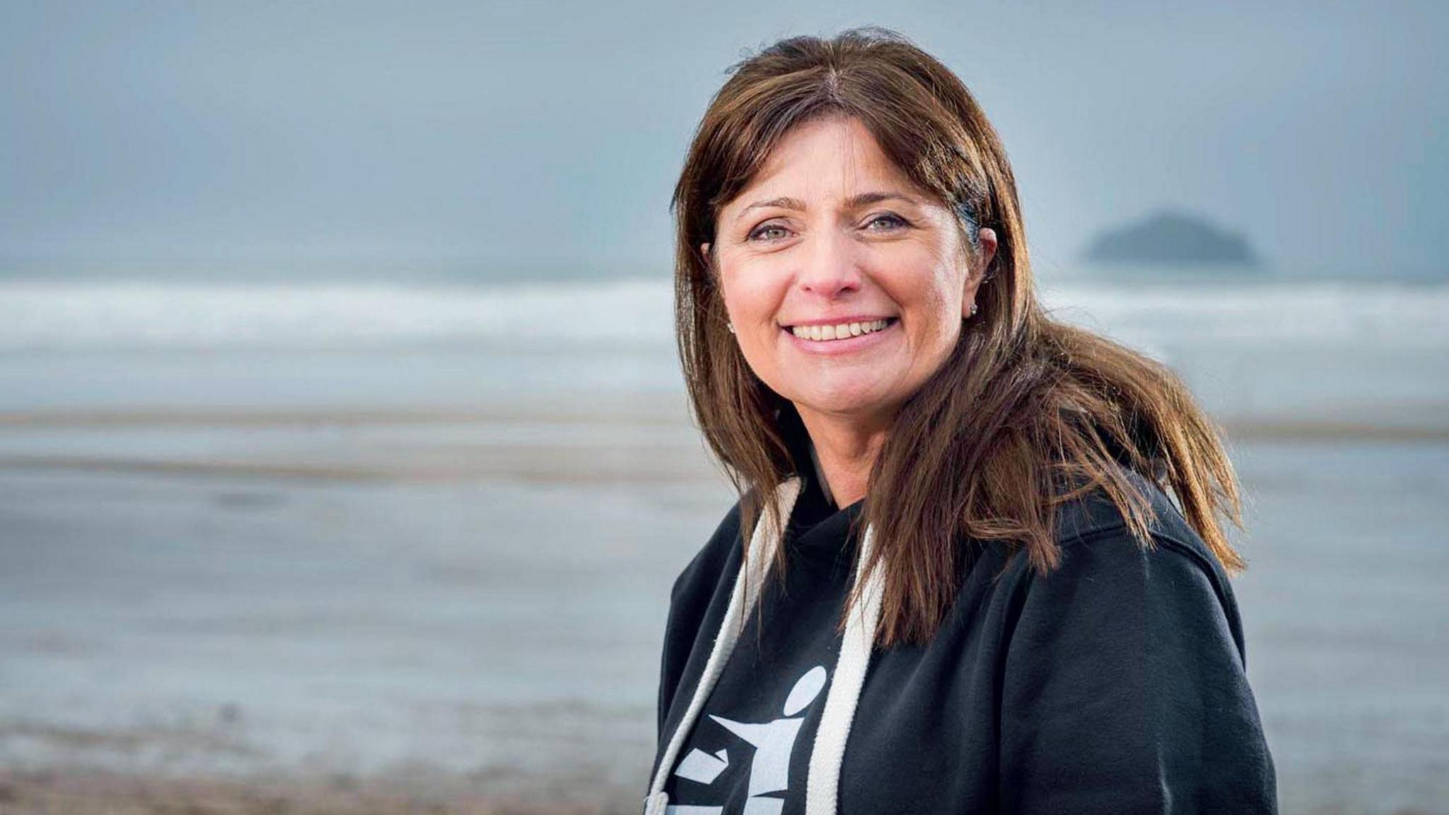 A brunette woman in a hoodie smiles at the camera from a beach.